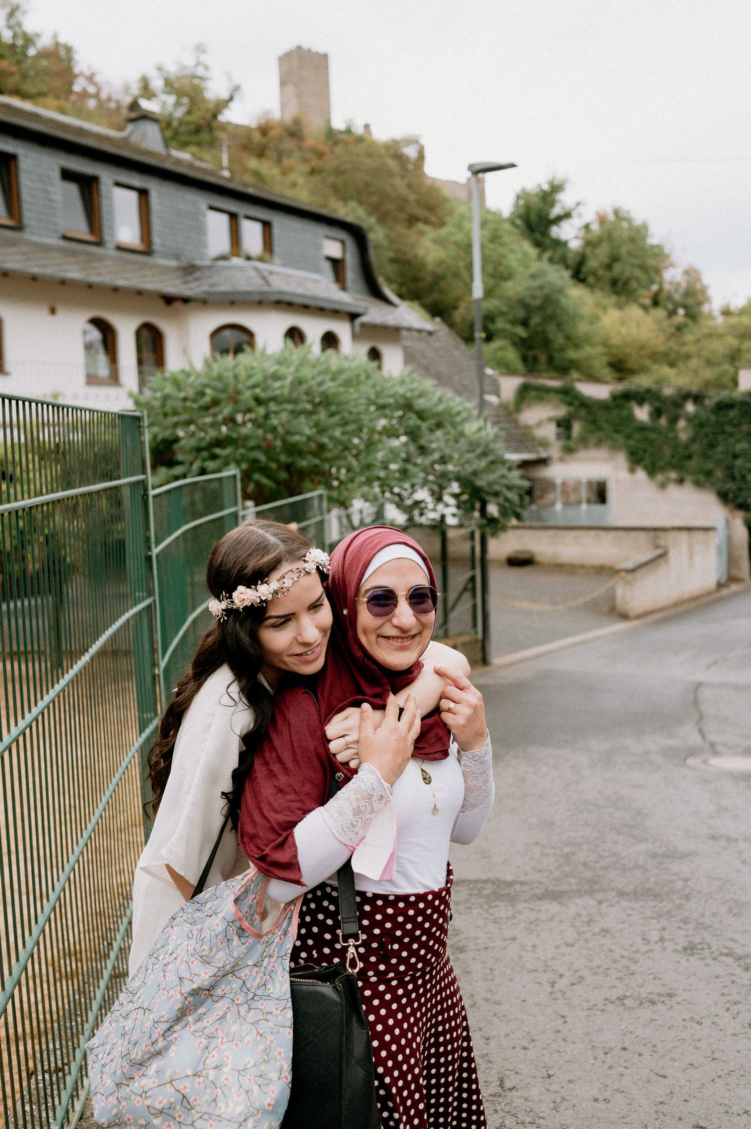Two friends embrace on a street with a castle tower visible on the hilltop behind them.