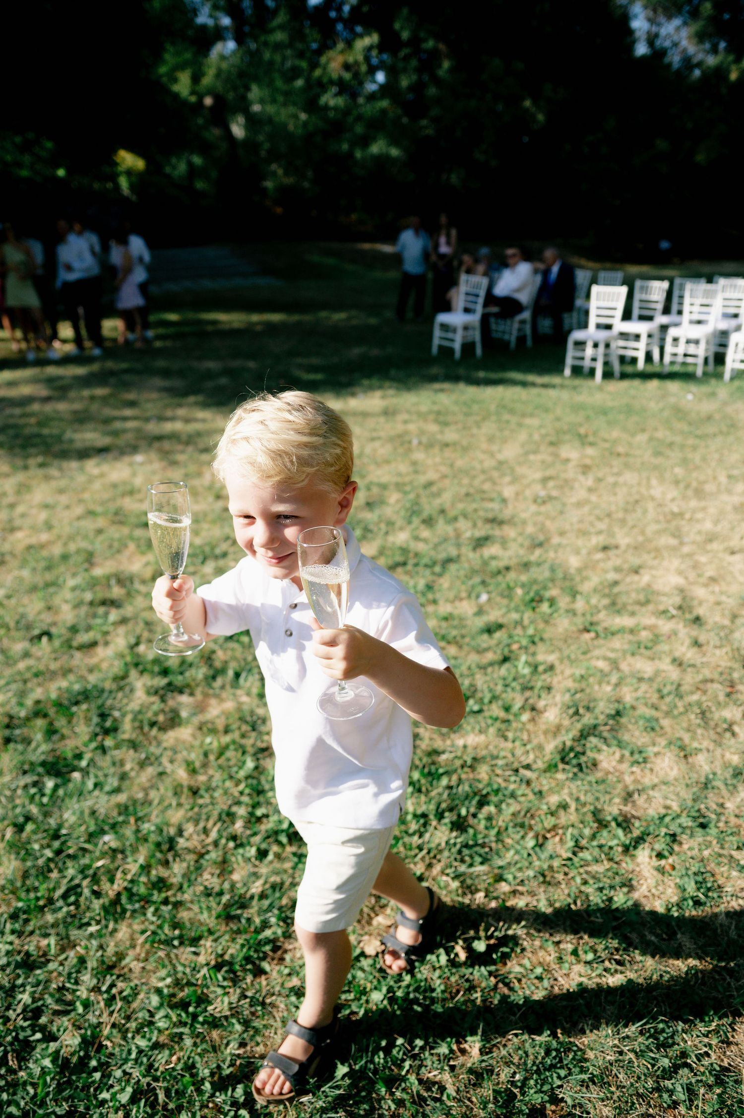 A toddler in white clothing plays in a sunny field near wedding ceremony chairs.