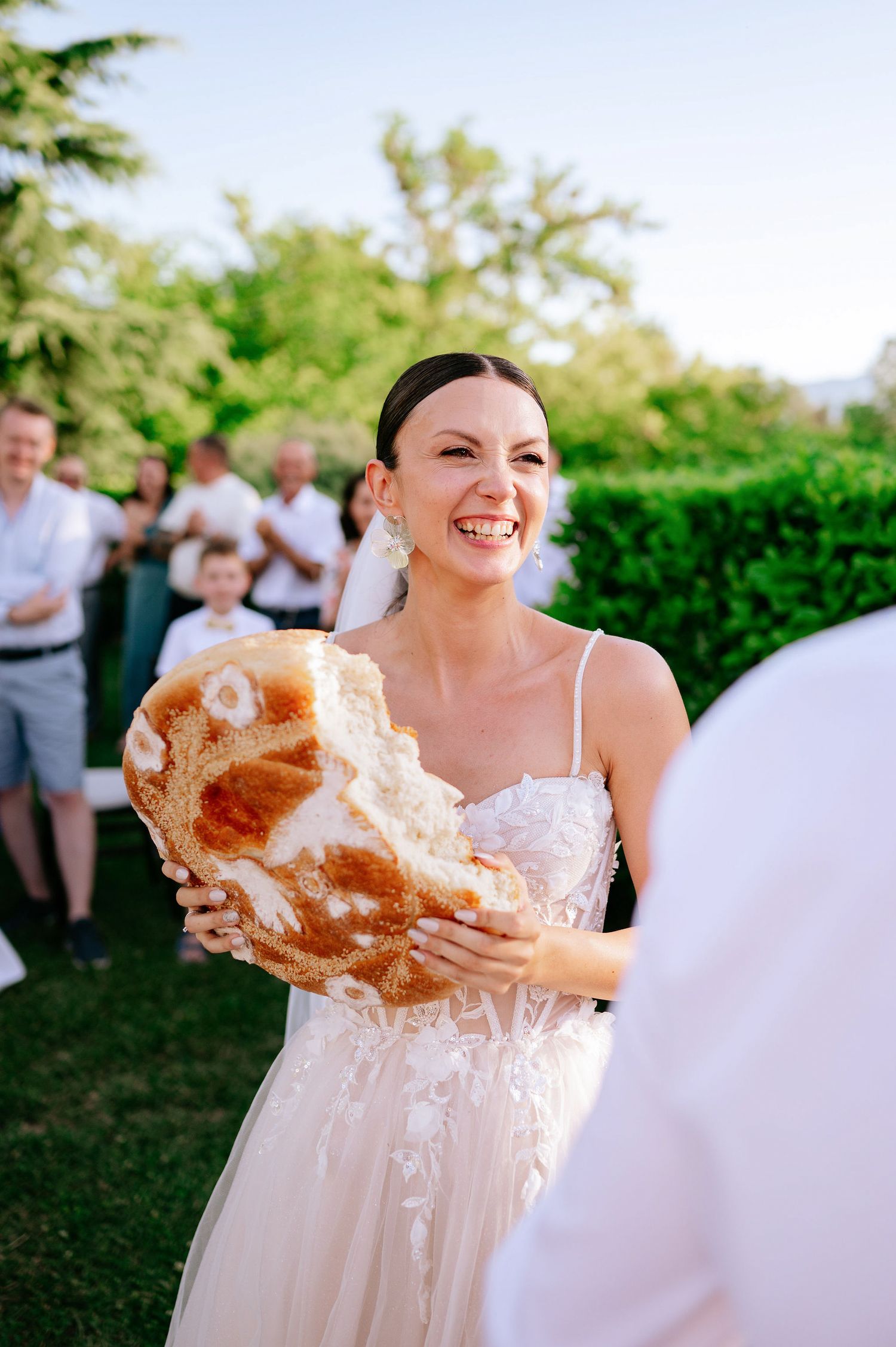 Person in a flowing white dress holds up a large loaf of bread outdoors.