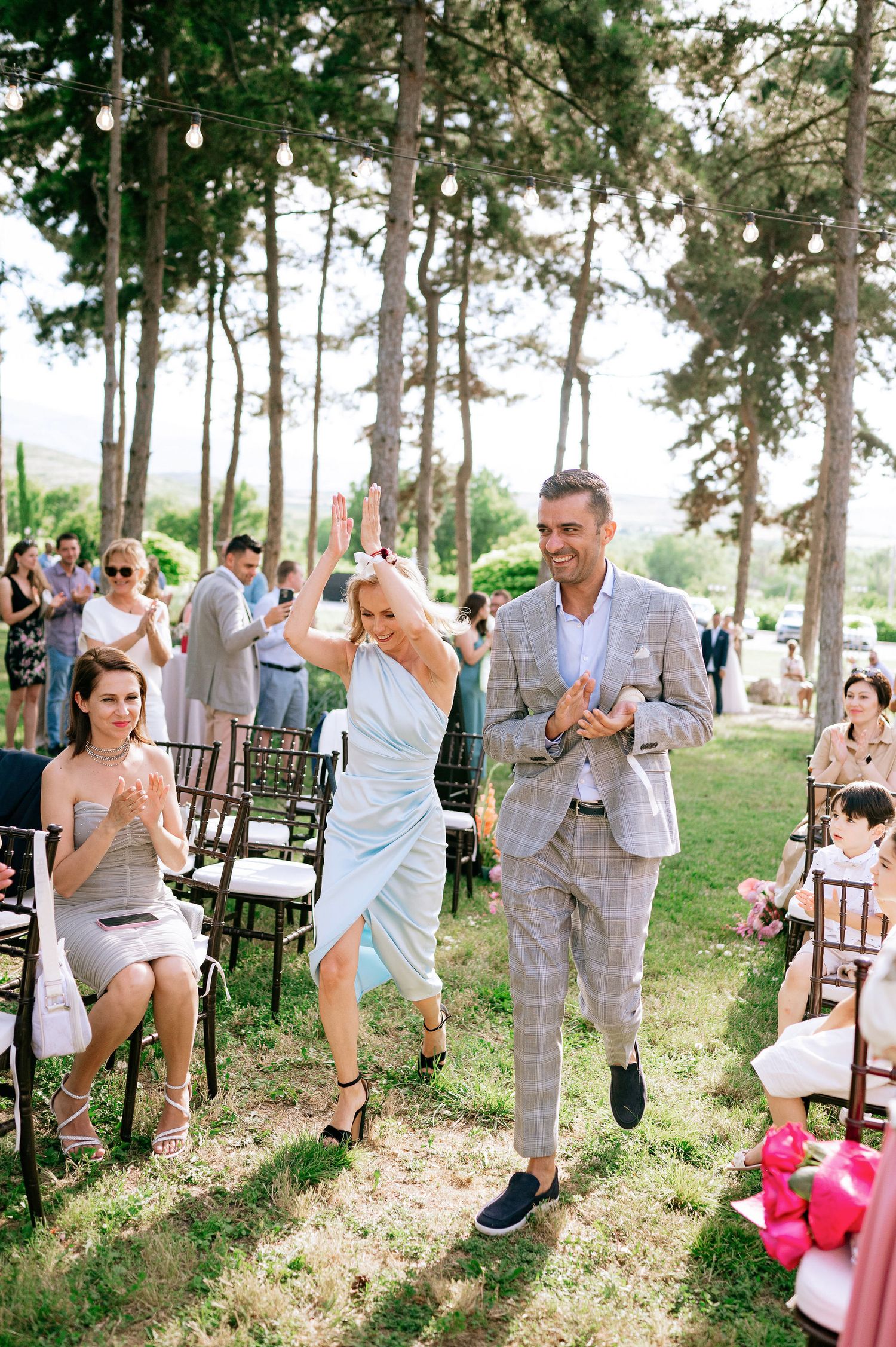 Wedding party members walking down outdoor aisle while guests applaud.