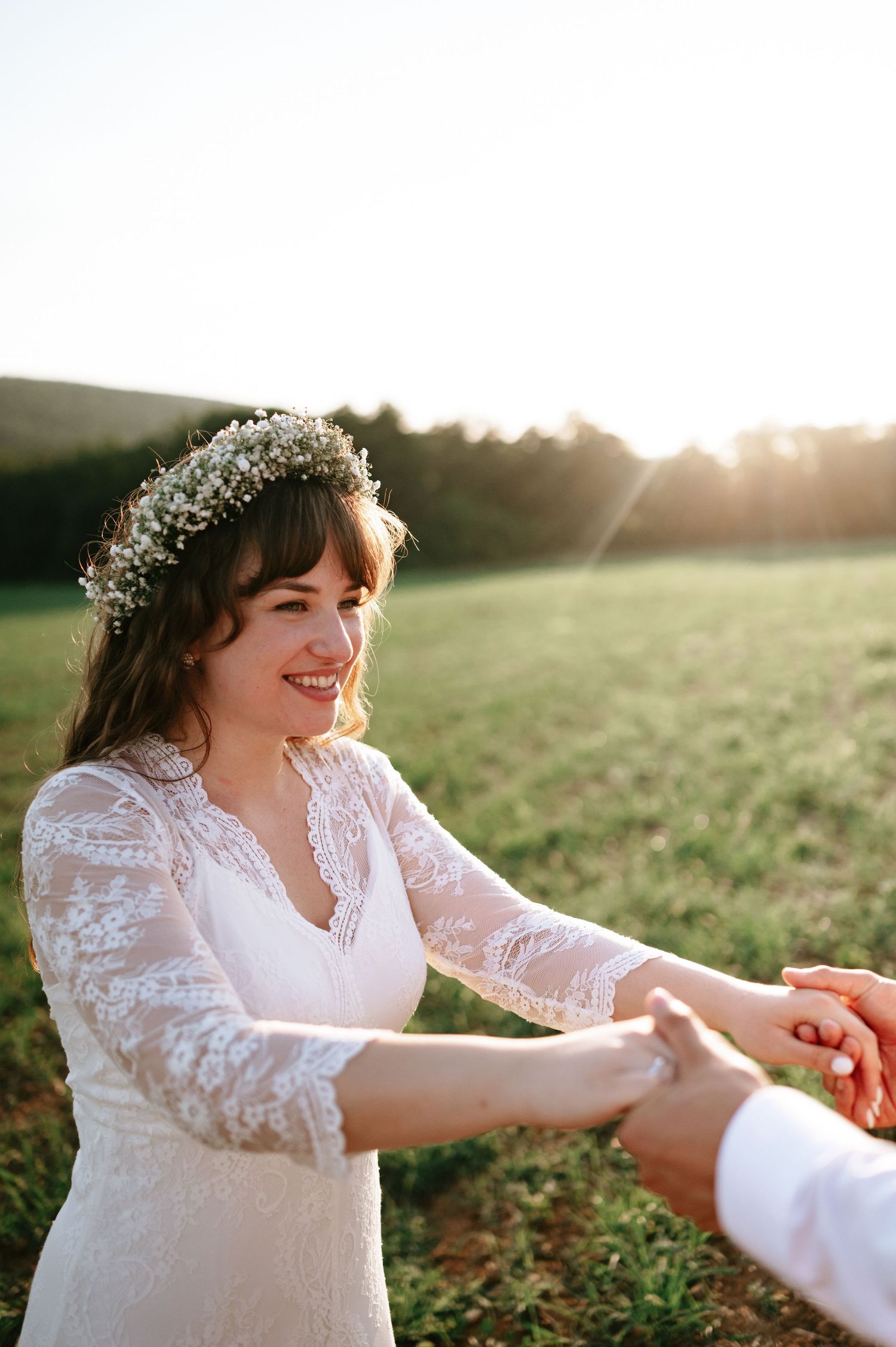 Romantic wedding couple holding hands in a sunlit field with the bride wearing a lace dress and floral crown.