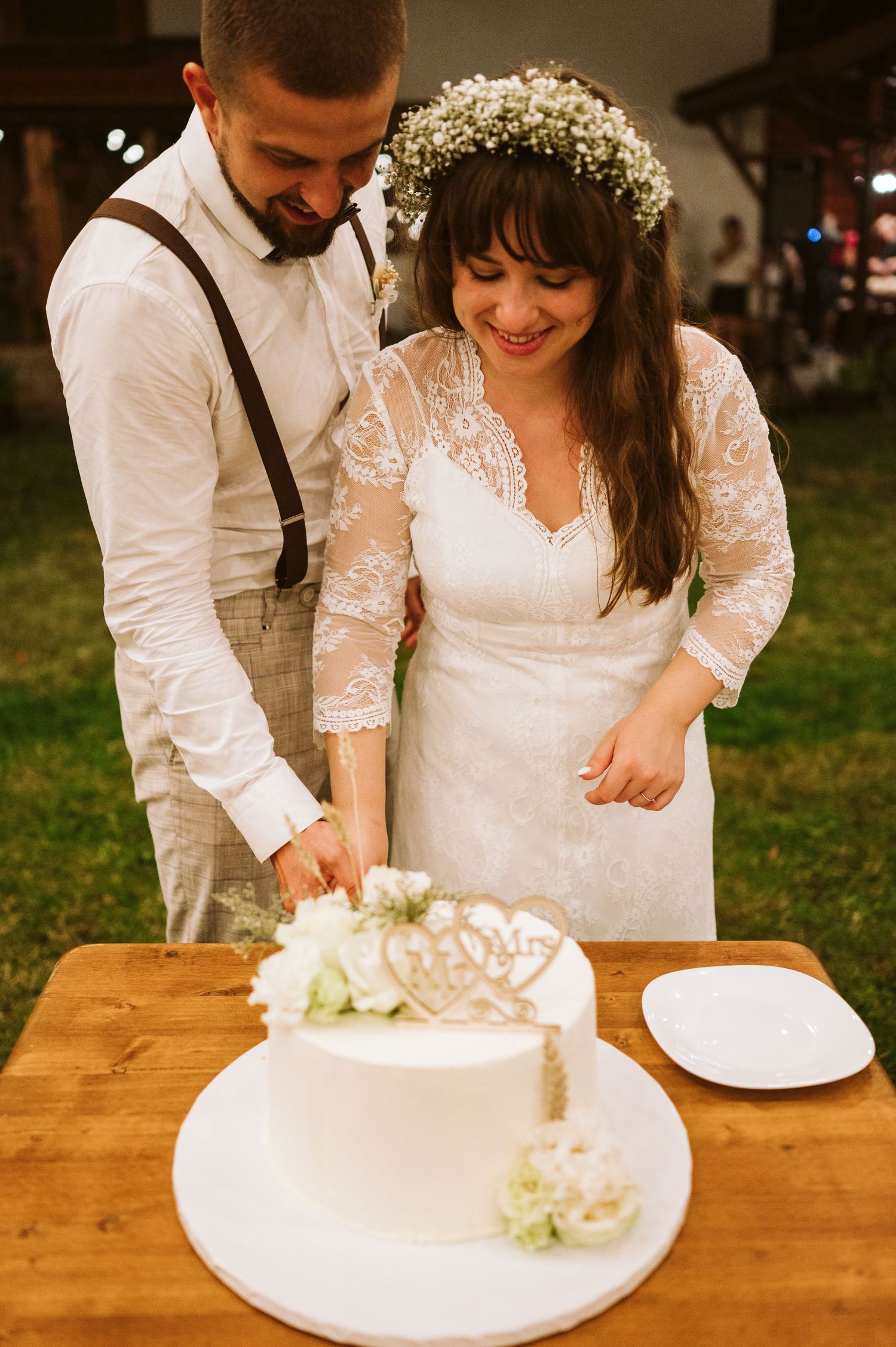 Newlyweds cut into their elegant white wedding cake together on a rustic wooden table.