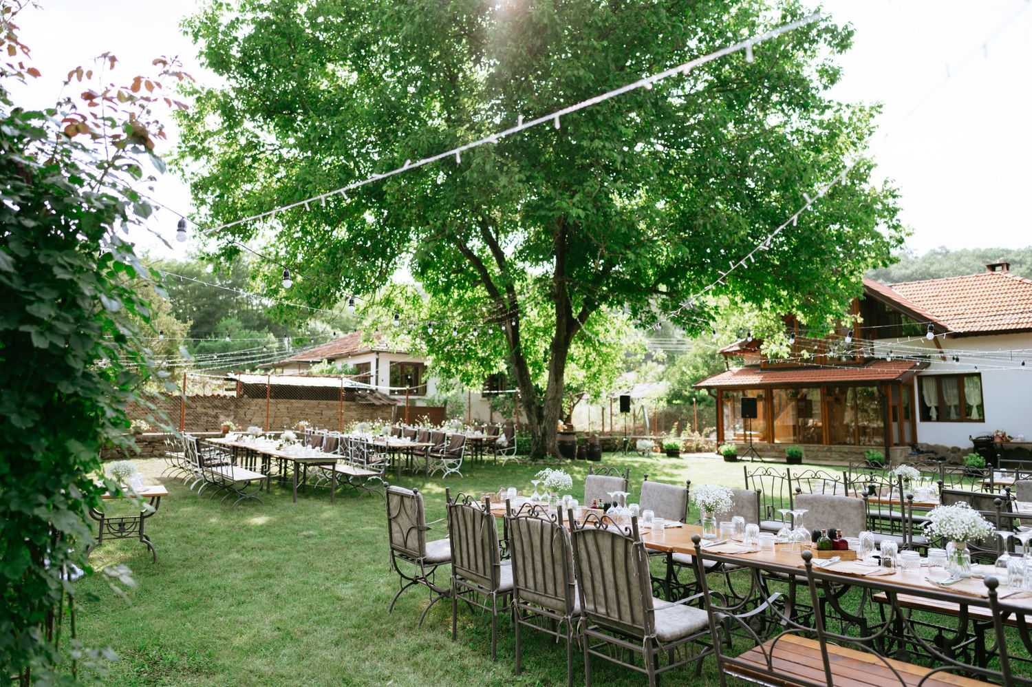 Outdoor wedding reception setup with wooden tables and chairs under string lights and large trees.