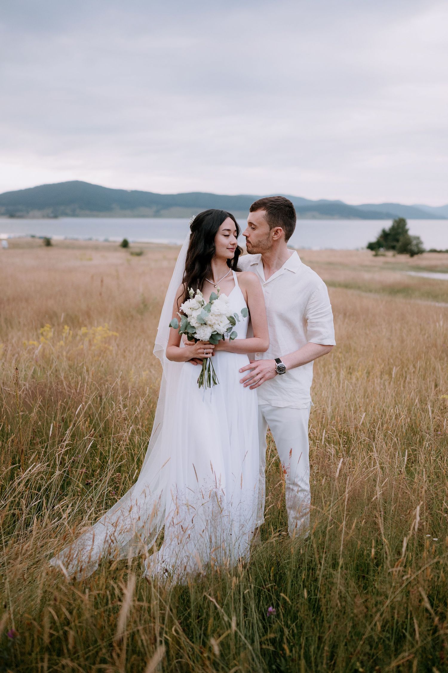 Wedding couple shares intimate moments in rustic field setting with mountain backdrop.