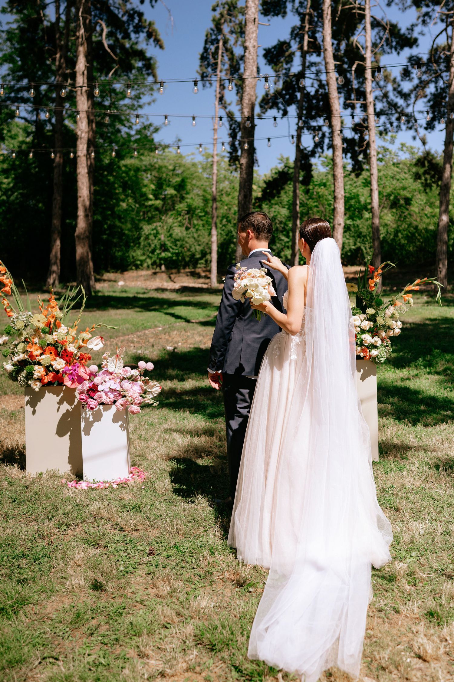 A wedding couple poses for romantic photos in a sunlit wooded outdoor setting with beautiful floral arrangements.