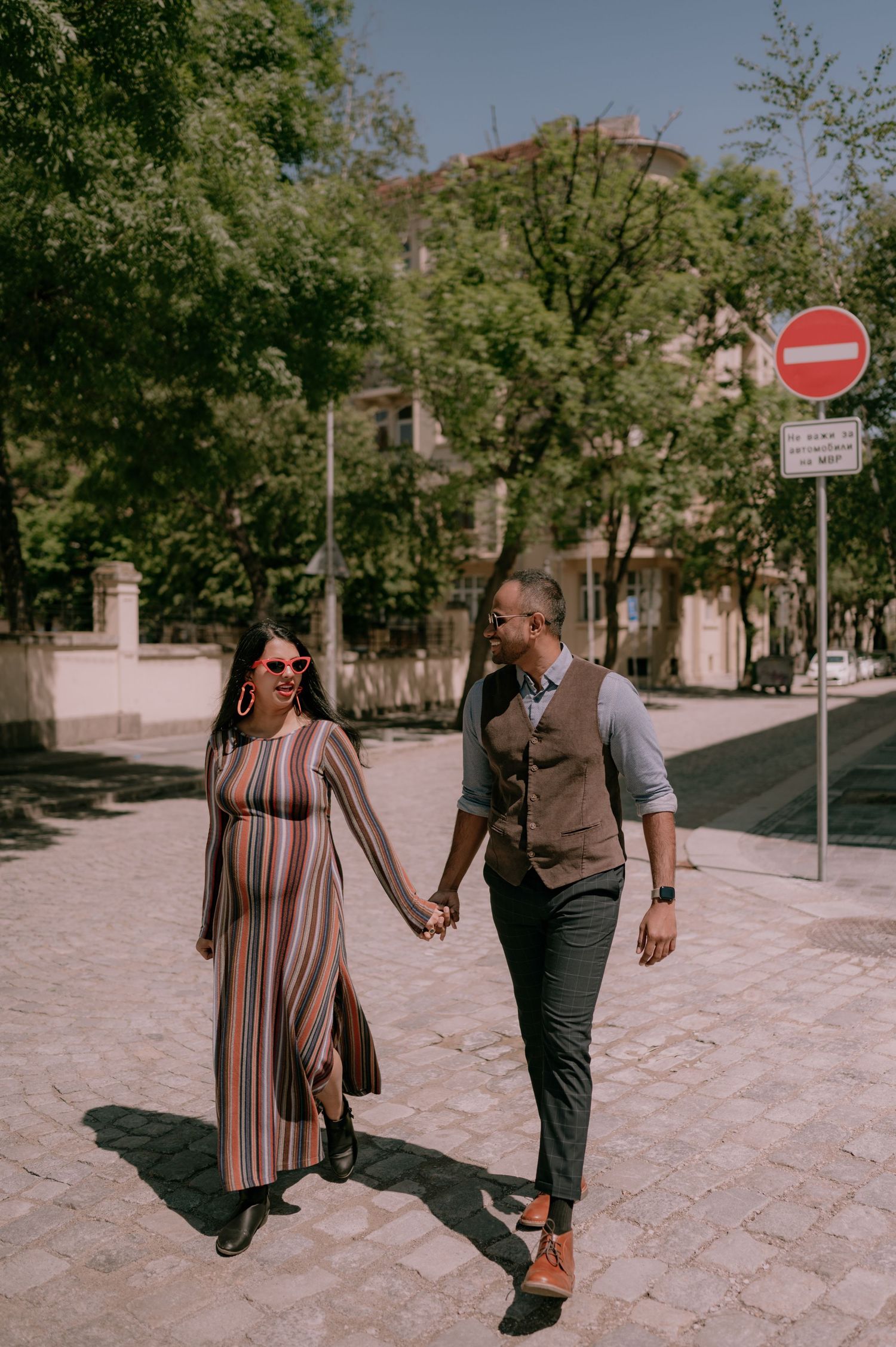 Pregnant woman in striped dress holds hands with man in vest walking on sunny cobblestone street.