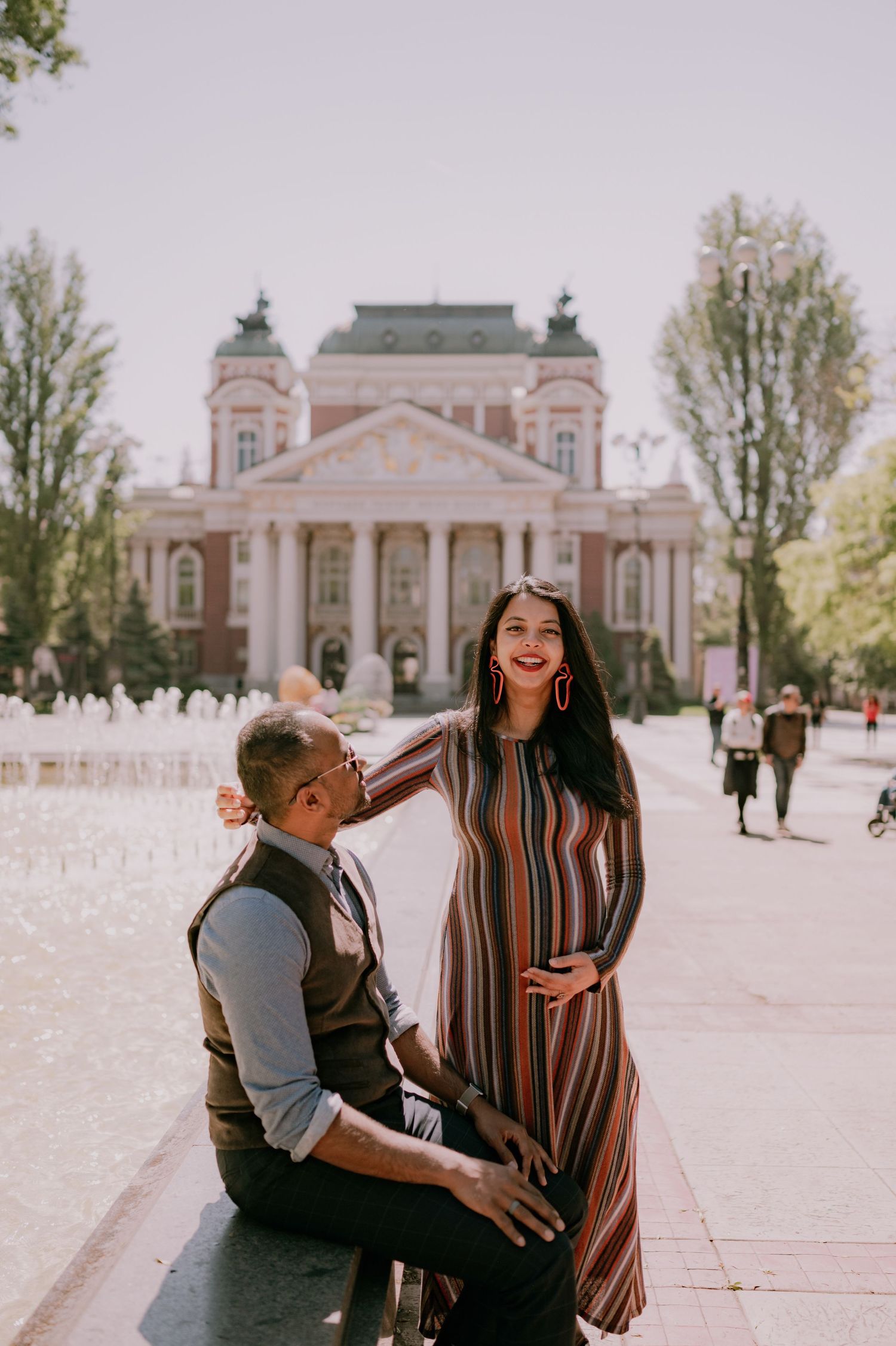 Couple posing in front of Ivan Vazov National Theatre in Sofia, Bulgaria, woman in striped dress smiling at seated man.