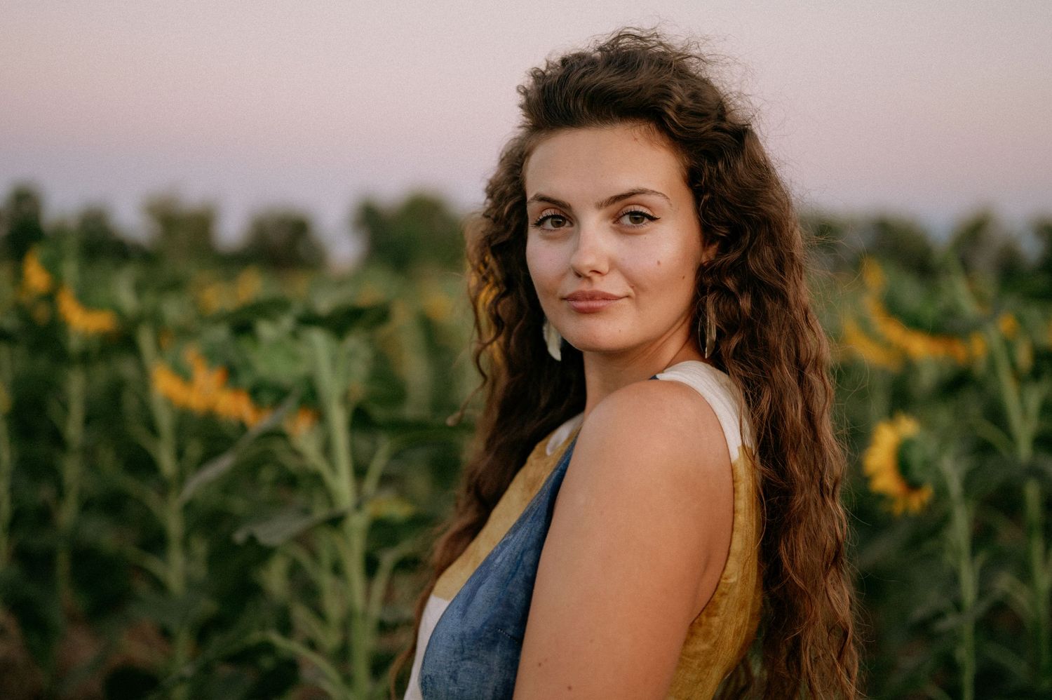 Young woman with curly hair poses in a sunflower field at sunset wearing a denim top.