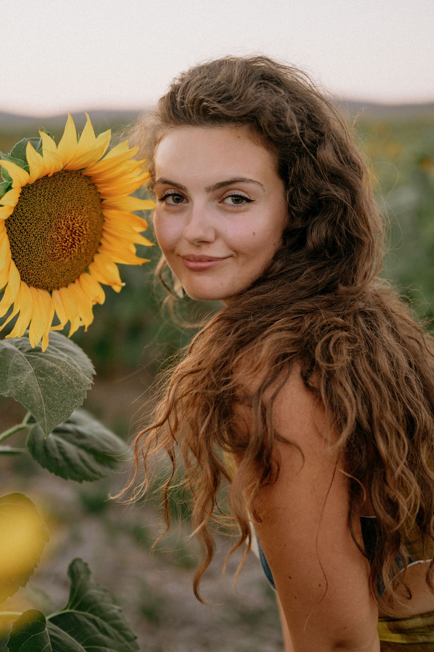 Young woman with curly hair smiling beside a bright yellow sunflower in a field at dusk.