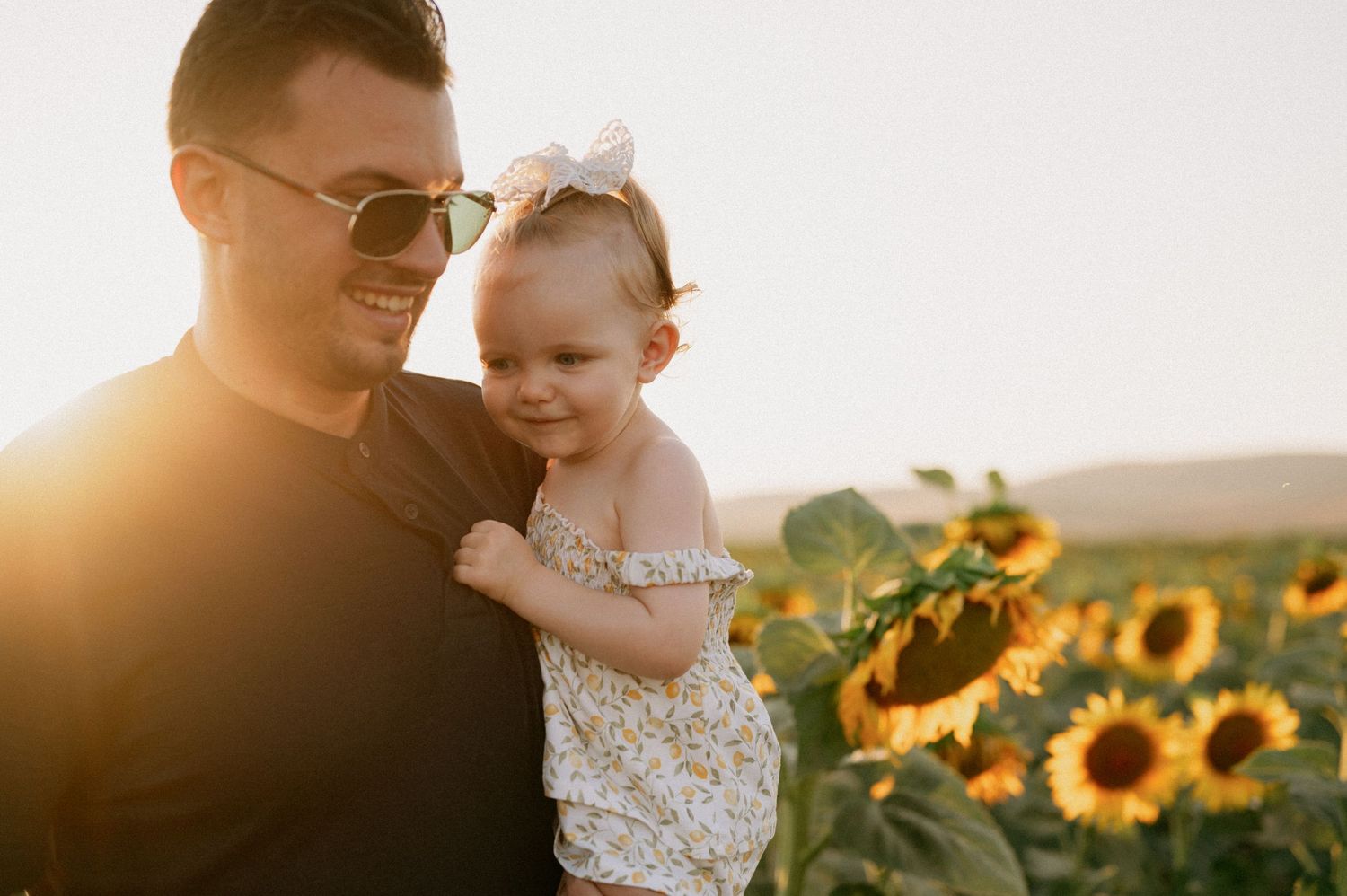 A father holds his toddler daughter in a sunflower field during golden hour sunset.