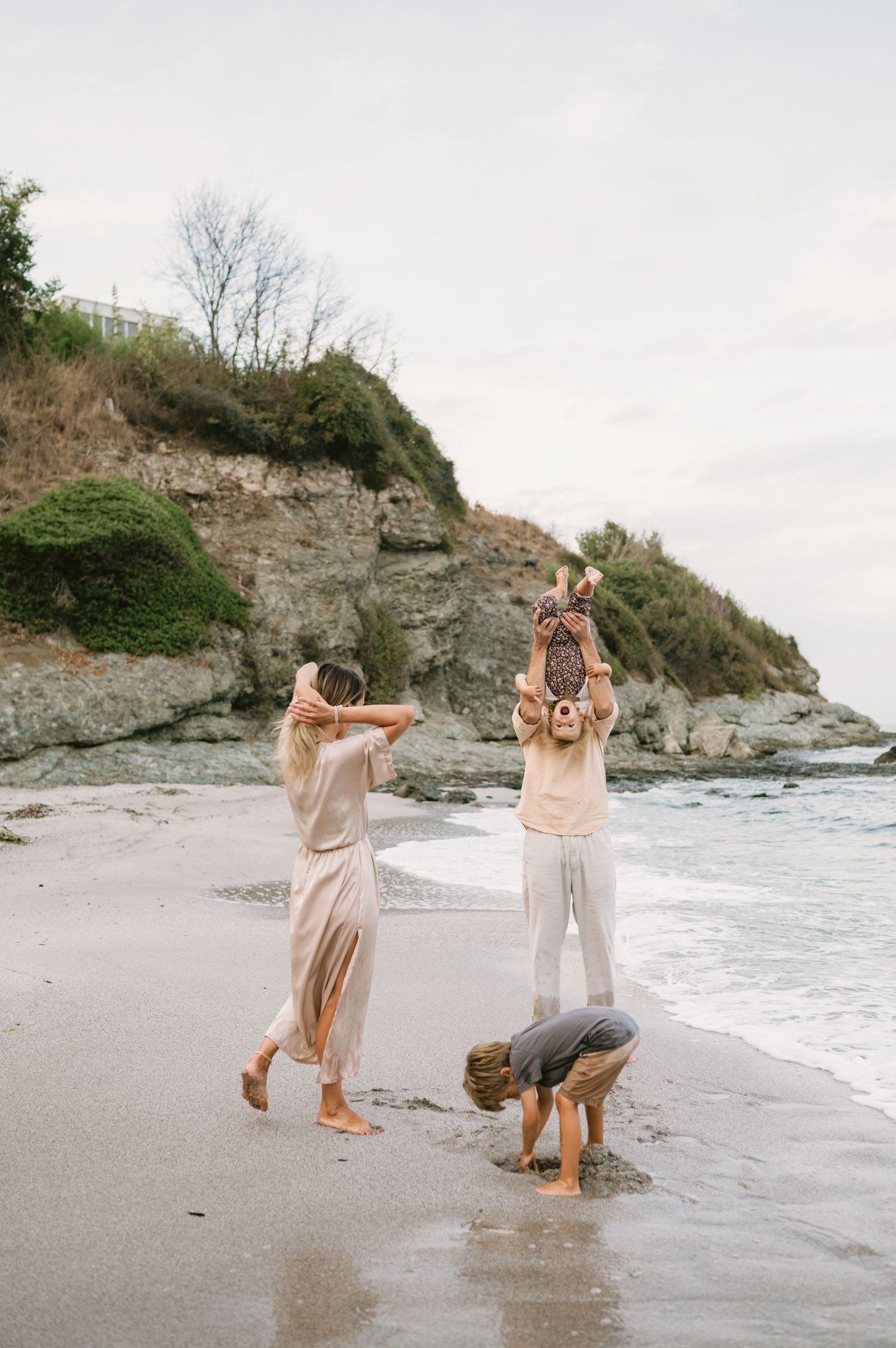 Family of three playing on a sandy beach with rocky cliffs, wearing neutral tones, as one person does a headstand.
