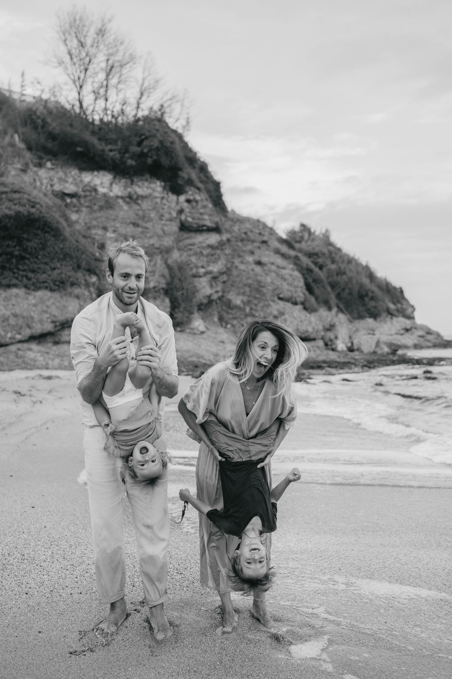 Black and white photo of a joyful family on a beach, parents playfully holding children upside down near rocky cliffs.