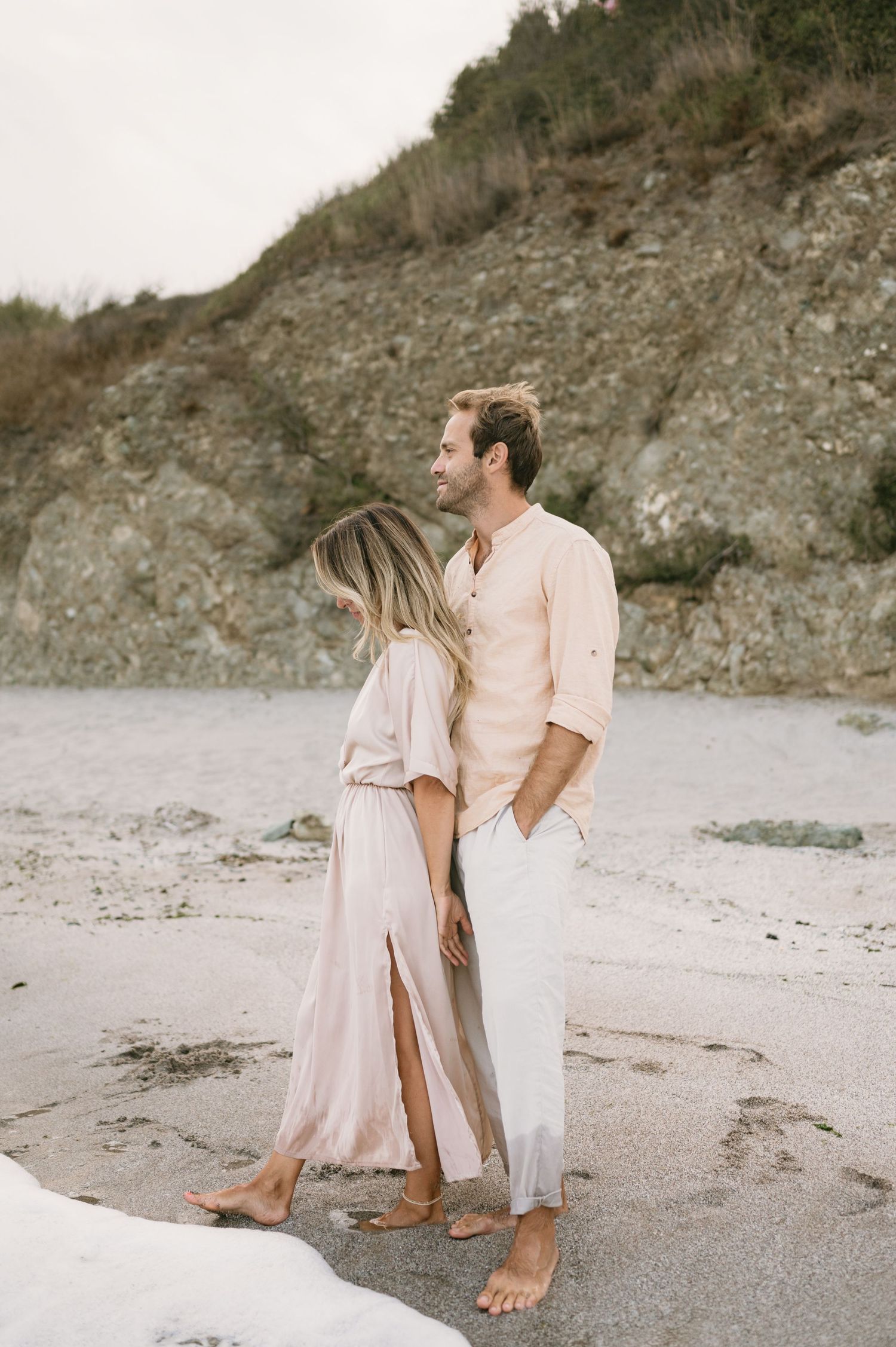 A couple in matching blush outfits share a romantic moment on a sandy beach with rocky cliffs behind them.