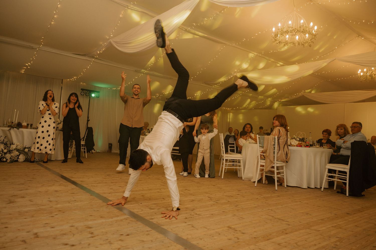 Person performing a one-handed handstand at an elegant wedding reception with guests watching in a decorated venue.