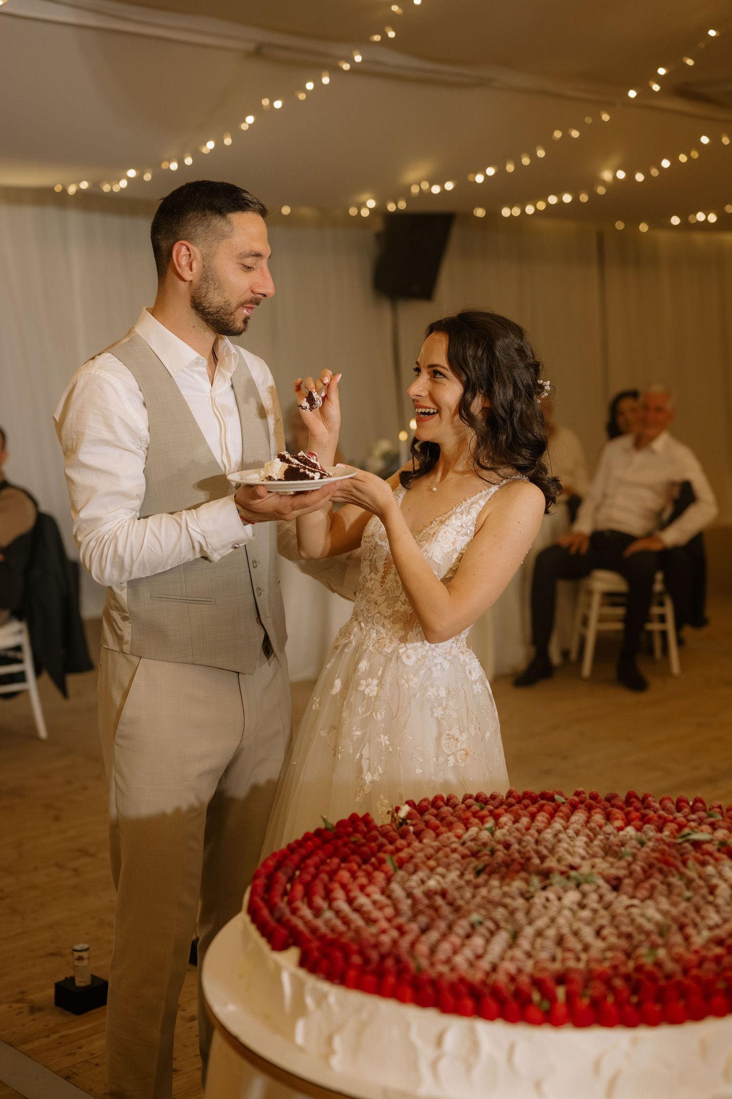 Bride and groom sharing wedding cake at reception, decorated with strawberries, under fairy lights.