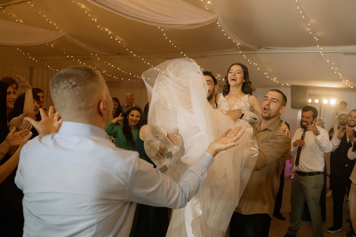 Bride lifted by guests during joyful wedding reception dance celebration with fairy lights backdrop.