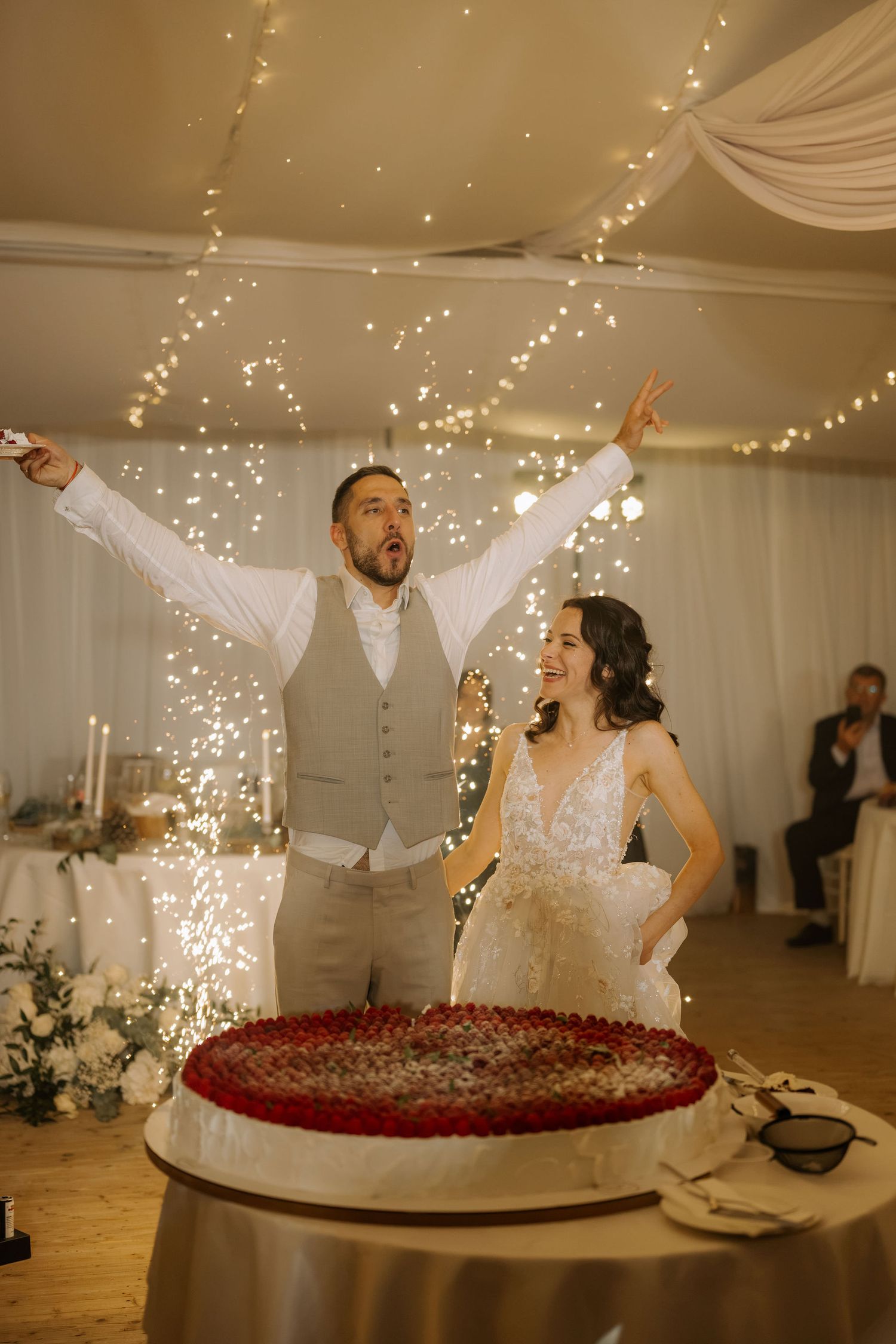 Bride and groom celebrating wedding cake cutting with sparklers under fairy lights in elegant reception venue.