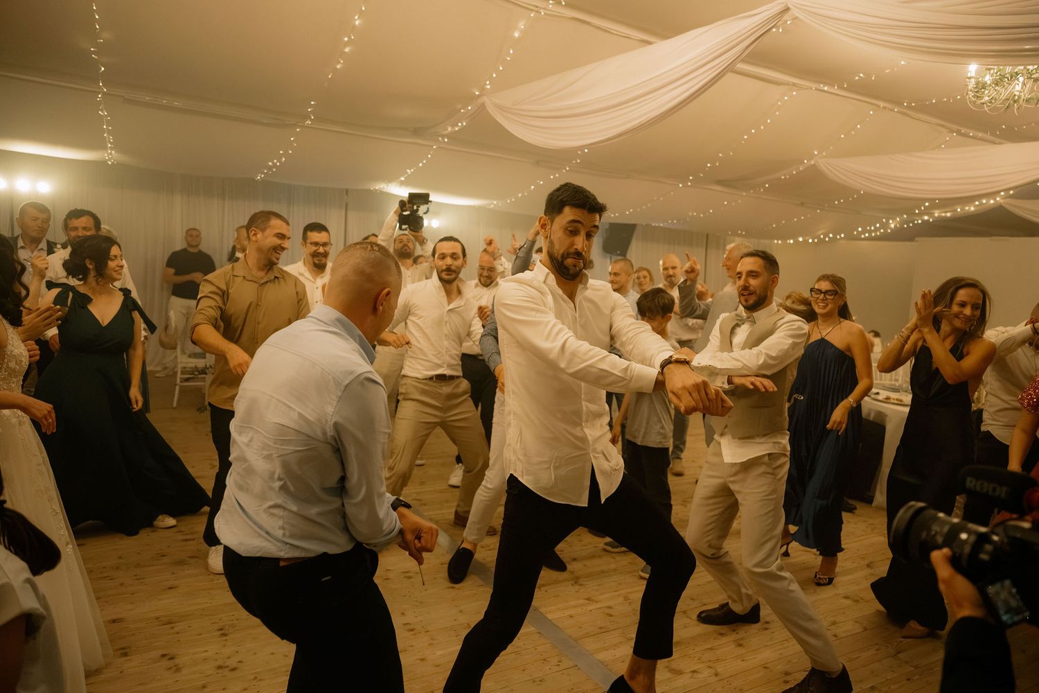 Men dancing energetically at a wedding reception in a decorated tent with string lights and guests watching.