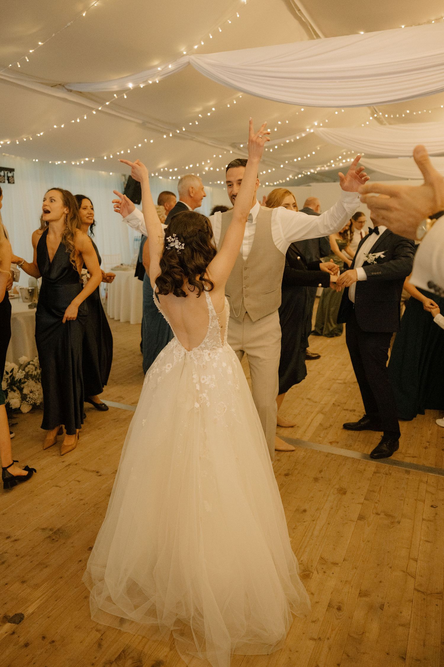 Bride and groom dancing joyfully at wedding reception in elegant tent venue with fairy lights and guests celebrating.