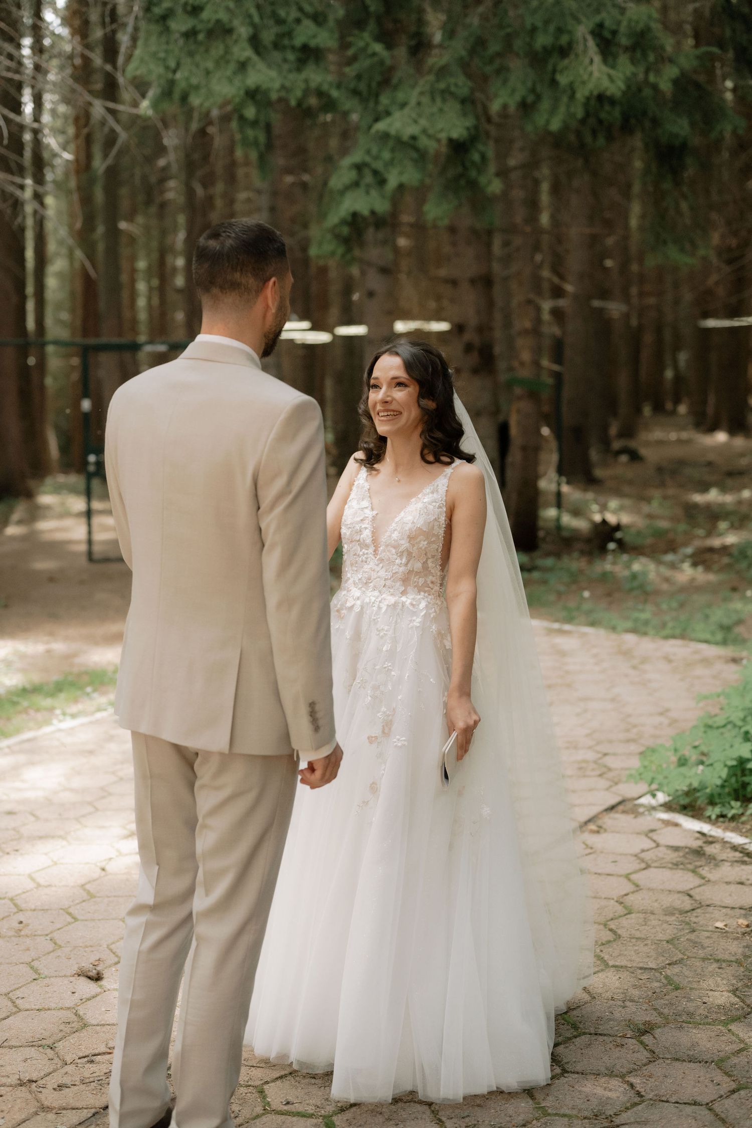 Bride in white lace gown and veil holds hands with groom in beige suit during outdoor forest wedding ceremony.