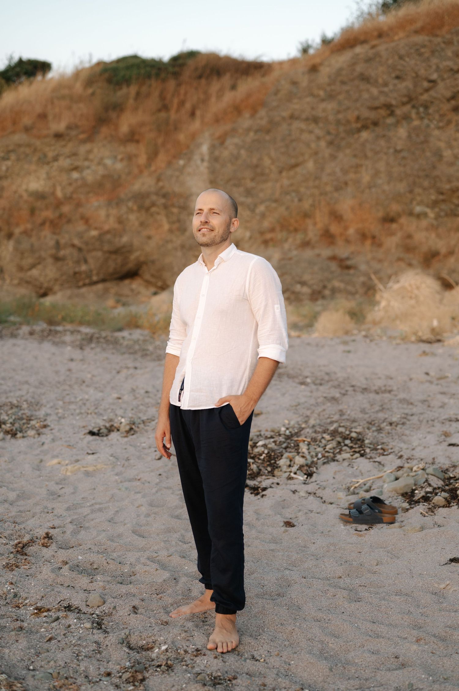 Person in pink shirt and dark shorts walks barefoot along rocky beach at sunset with cliffs in background.