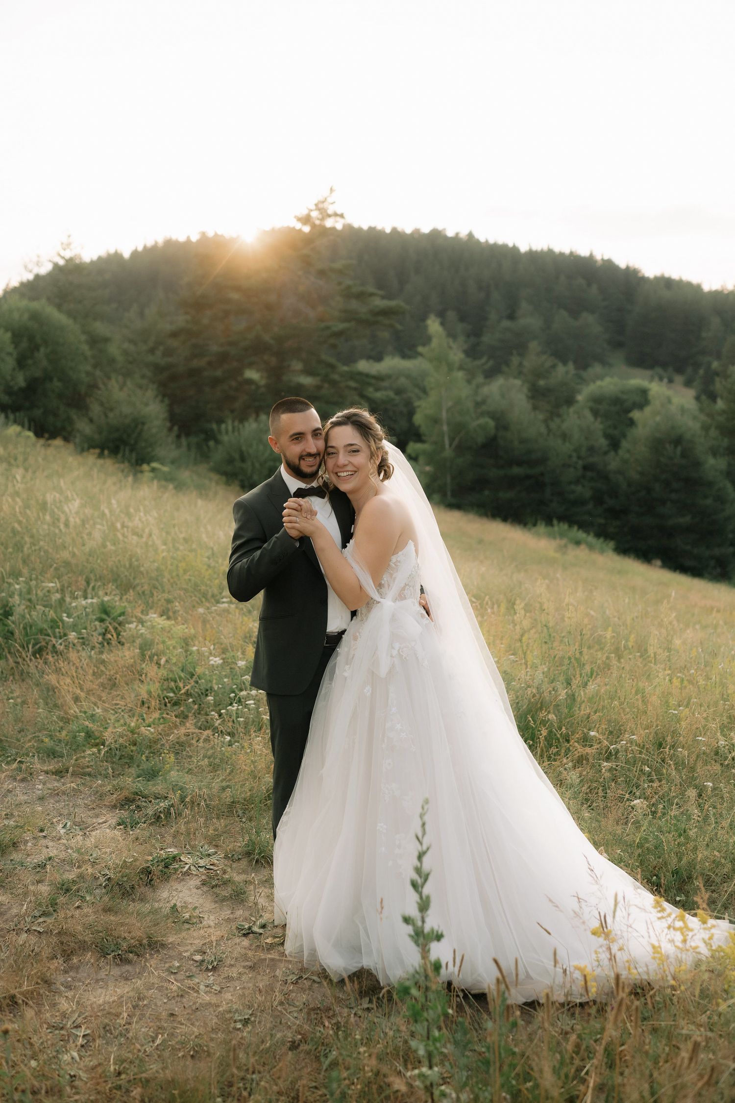 A couple in wedding attire embraces in a sunlit meadow with evergreen trees in the background during golden hour.