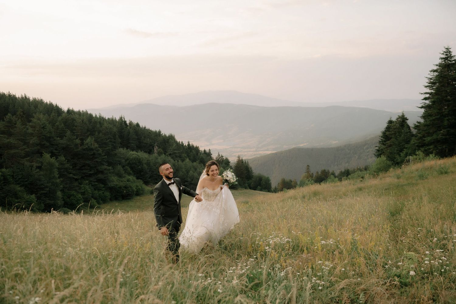 A couple walks through a misty mountain meadow during a romantic wedding photo session at sunset.