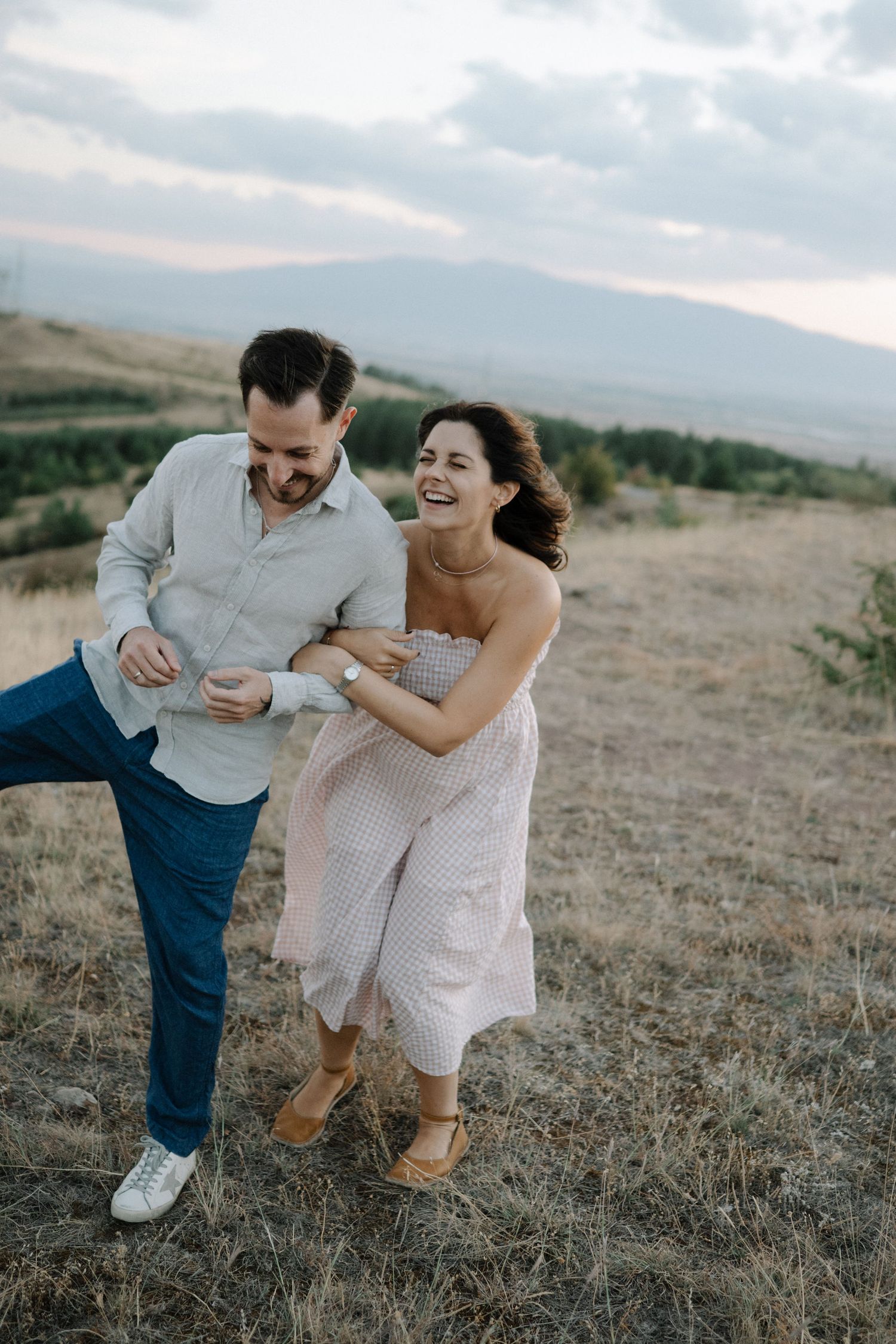 A couple shares a playful moment together while walking in a grassy field at sunset.
