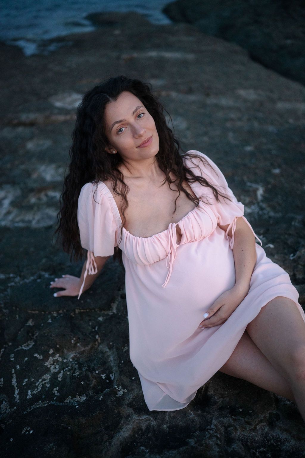 A person in a pink blouse poses on dark rocks during a moody outdoor photoshoot at dusk.