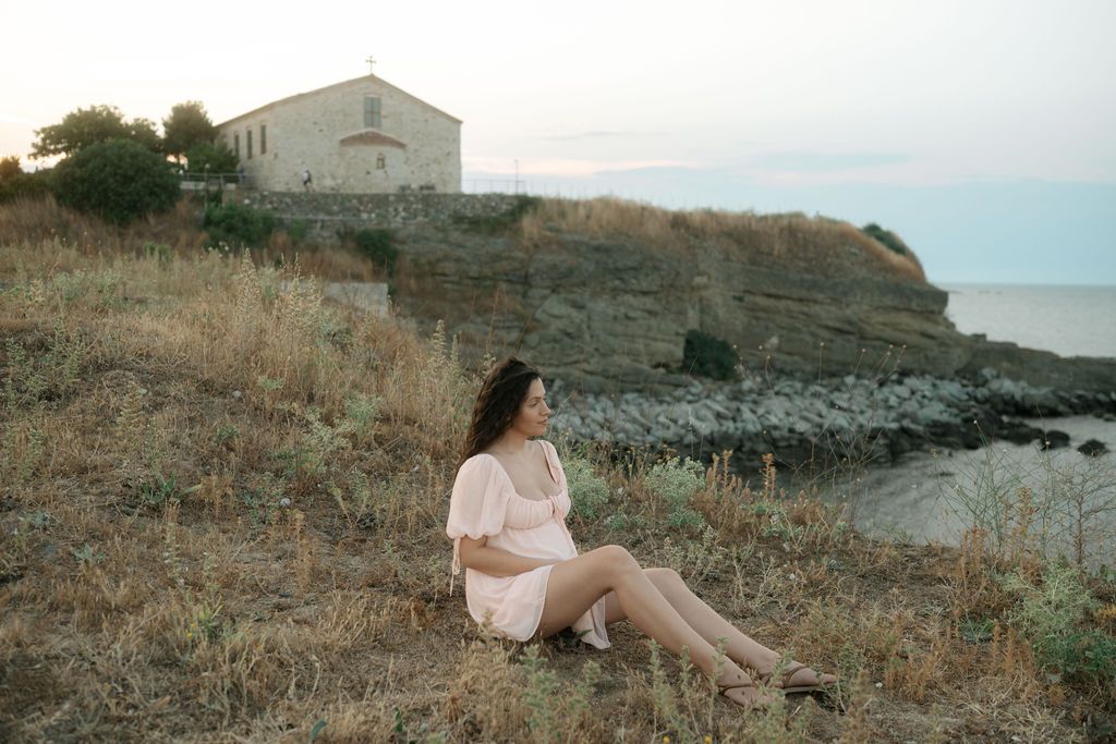 A person in a pink dress sits on a coastal hillside at dusk with a white Mediterranean building perched above the rocky shore.