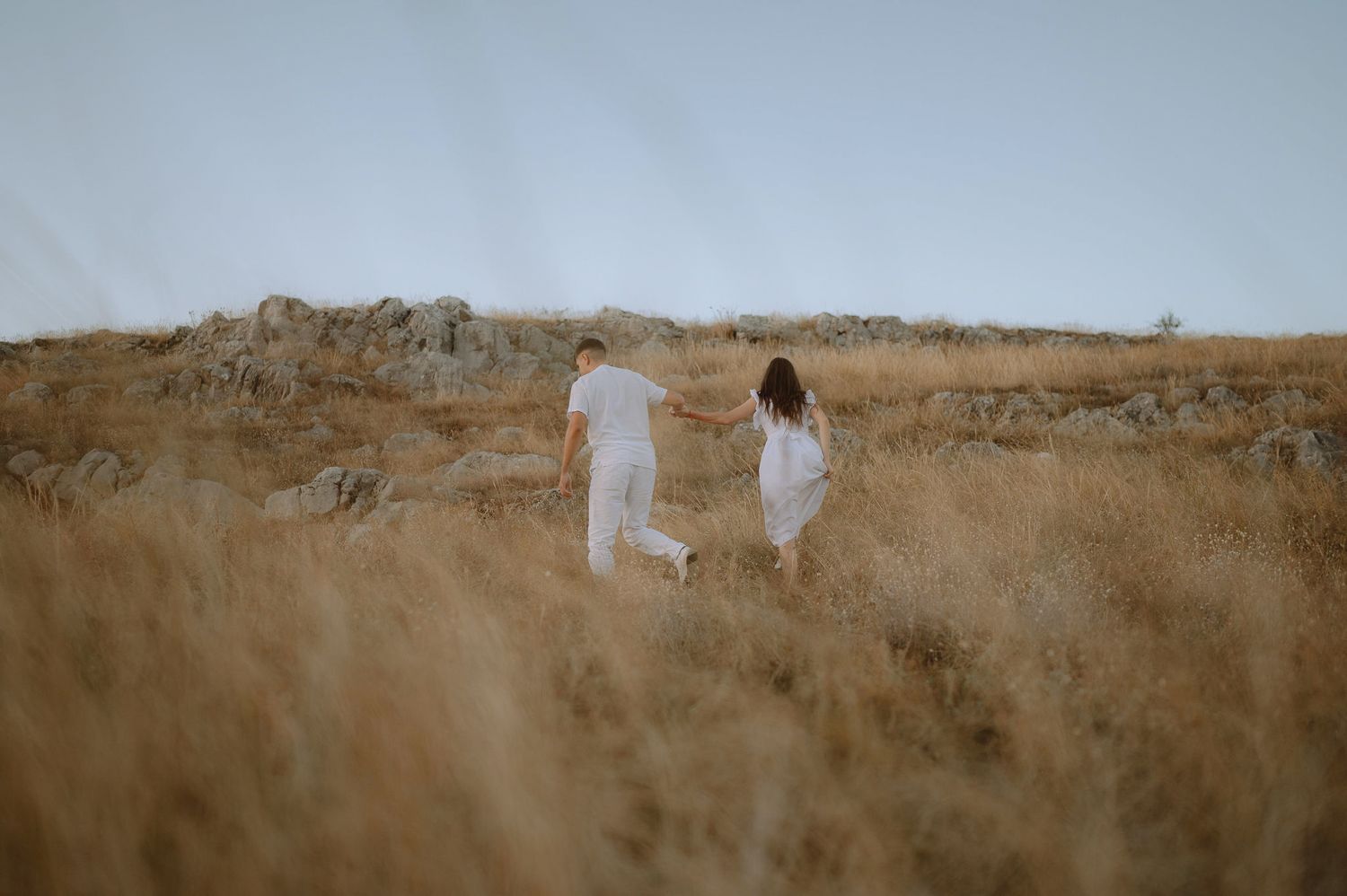 Two people in white dresses walk together through a golden field against a misty sky backdrop.