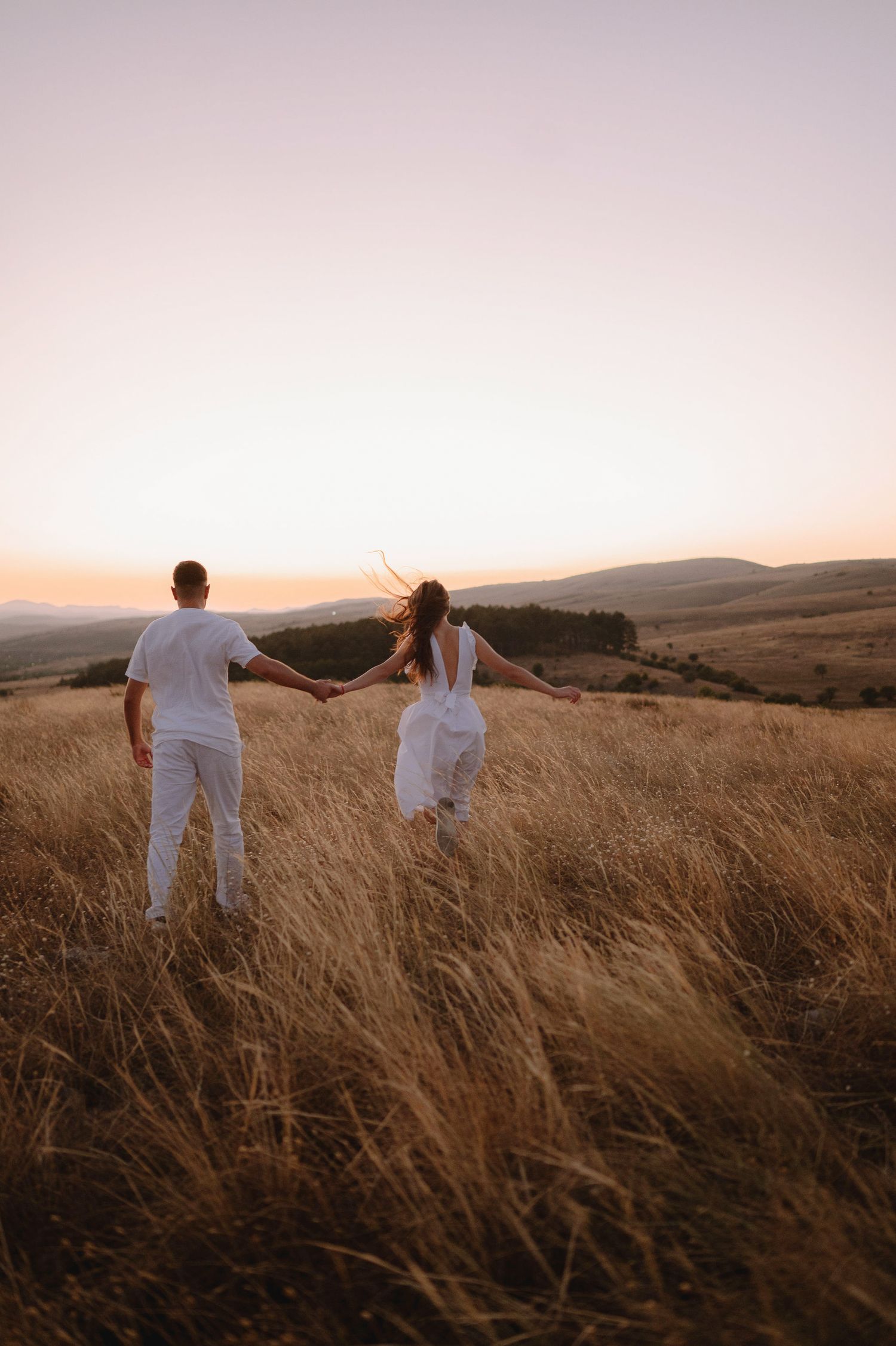 A couple in white outfits holding hands while standing in a golden field at sunset with rolling hills in background.