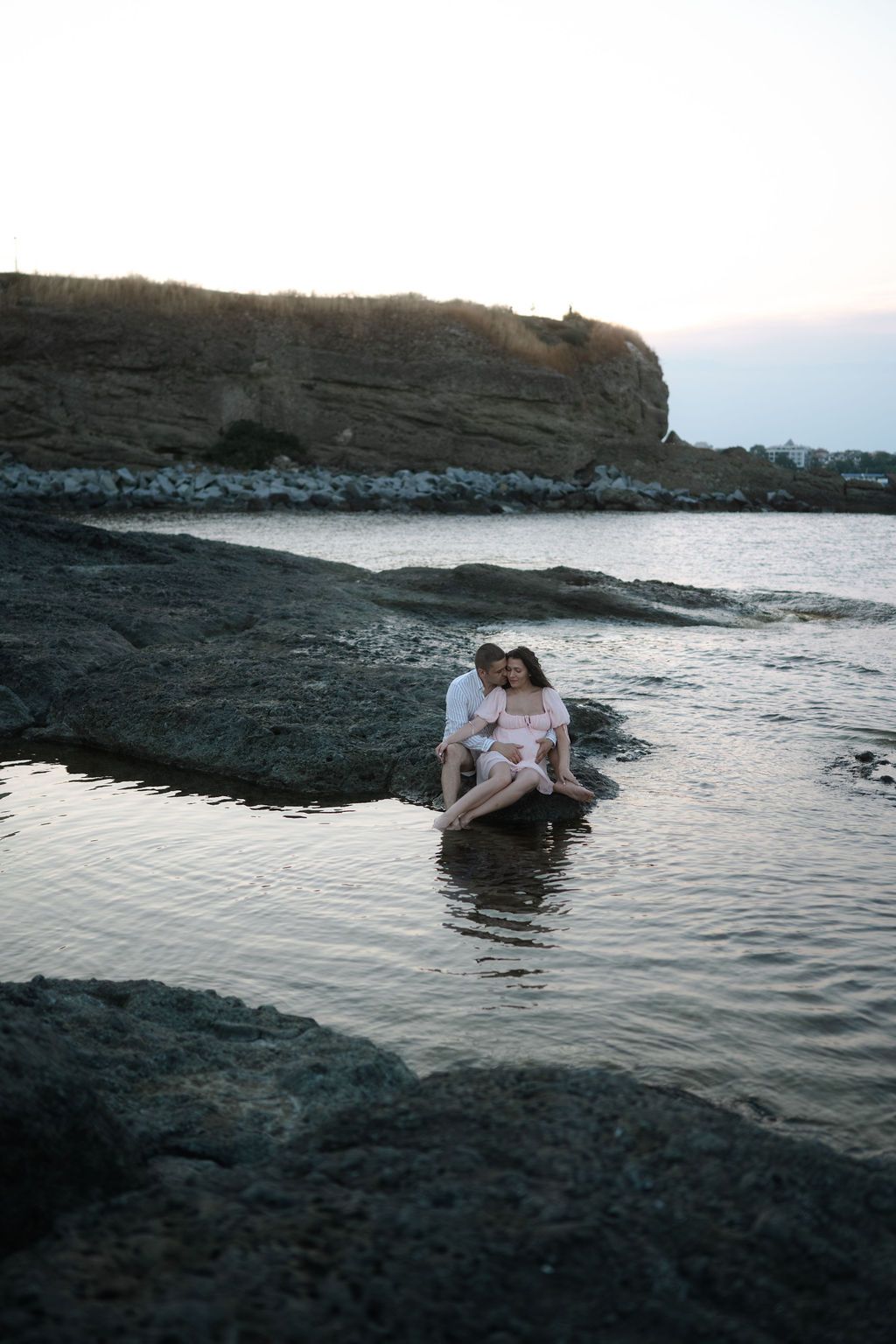 A couple shares an intimate moment on dark rocky shores at sunset as waves gently lap against the coastline.