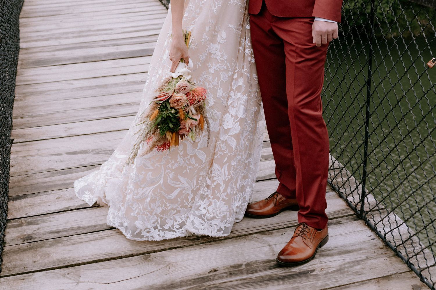 A couple in wedding attire stands on a rustic wooden deck, the bride in white lace and groom in burgundy suit.