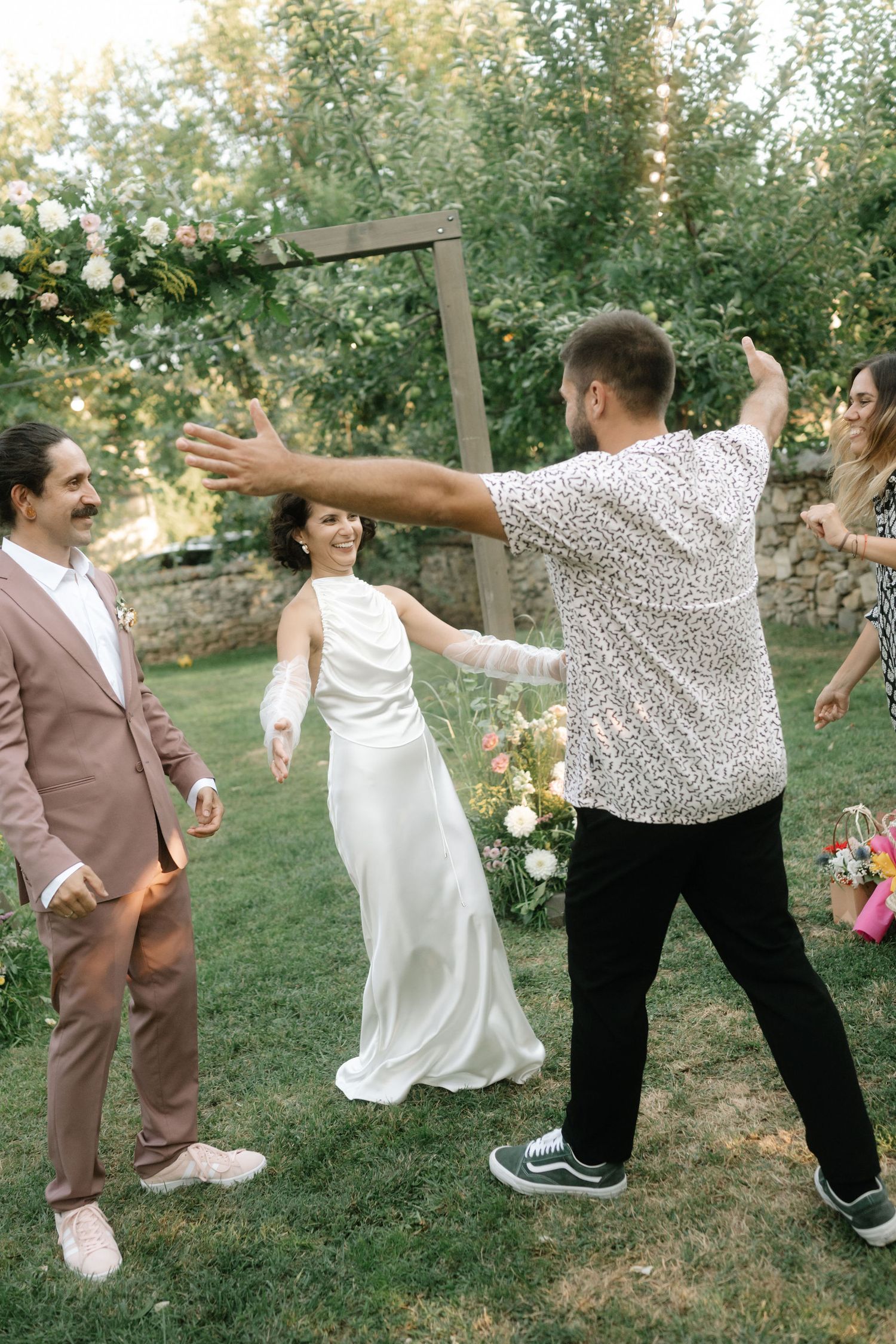 A wedding party performs a synchronized dance move in an outdoor garden setting with trees in the background.