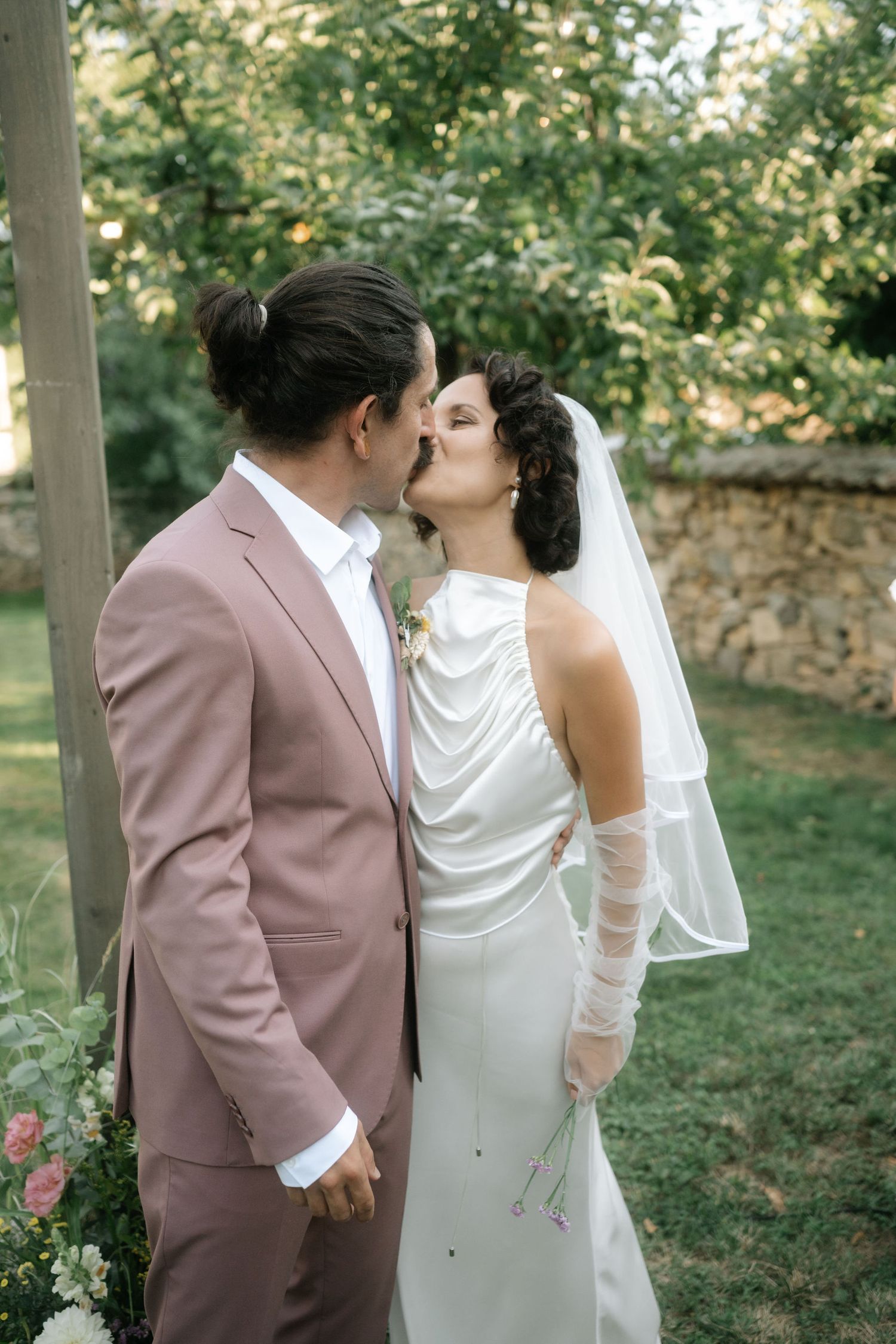 A romantic couple in wedding attire share intimate moments in a garden setting with stone walls and greenery.