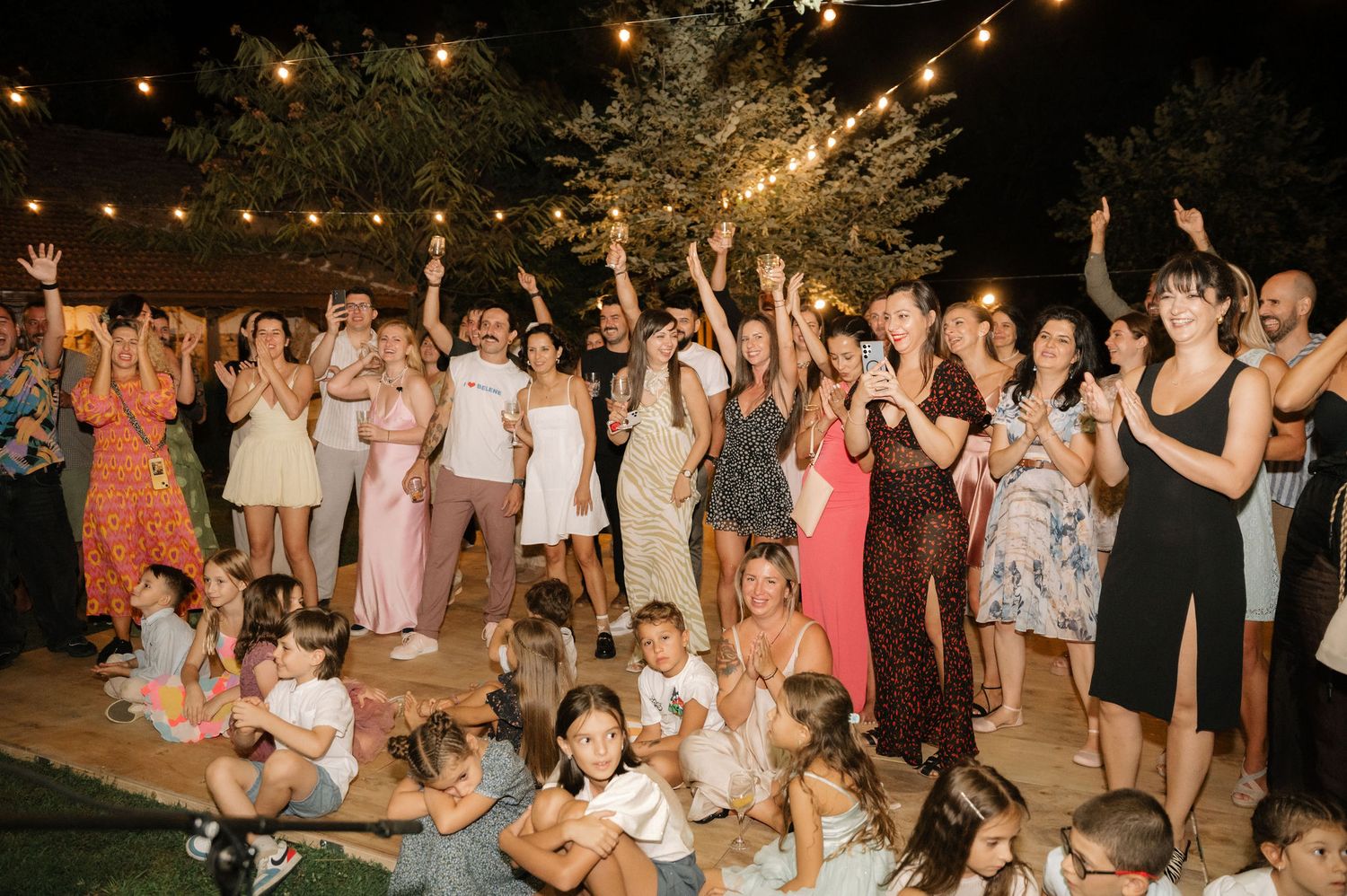 A large group of party guests dancing and celebrating outdoors under string lights at night.