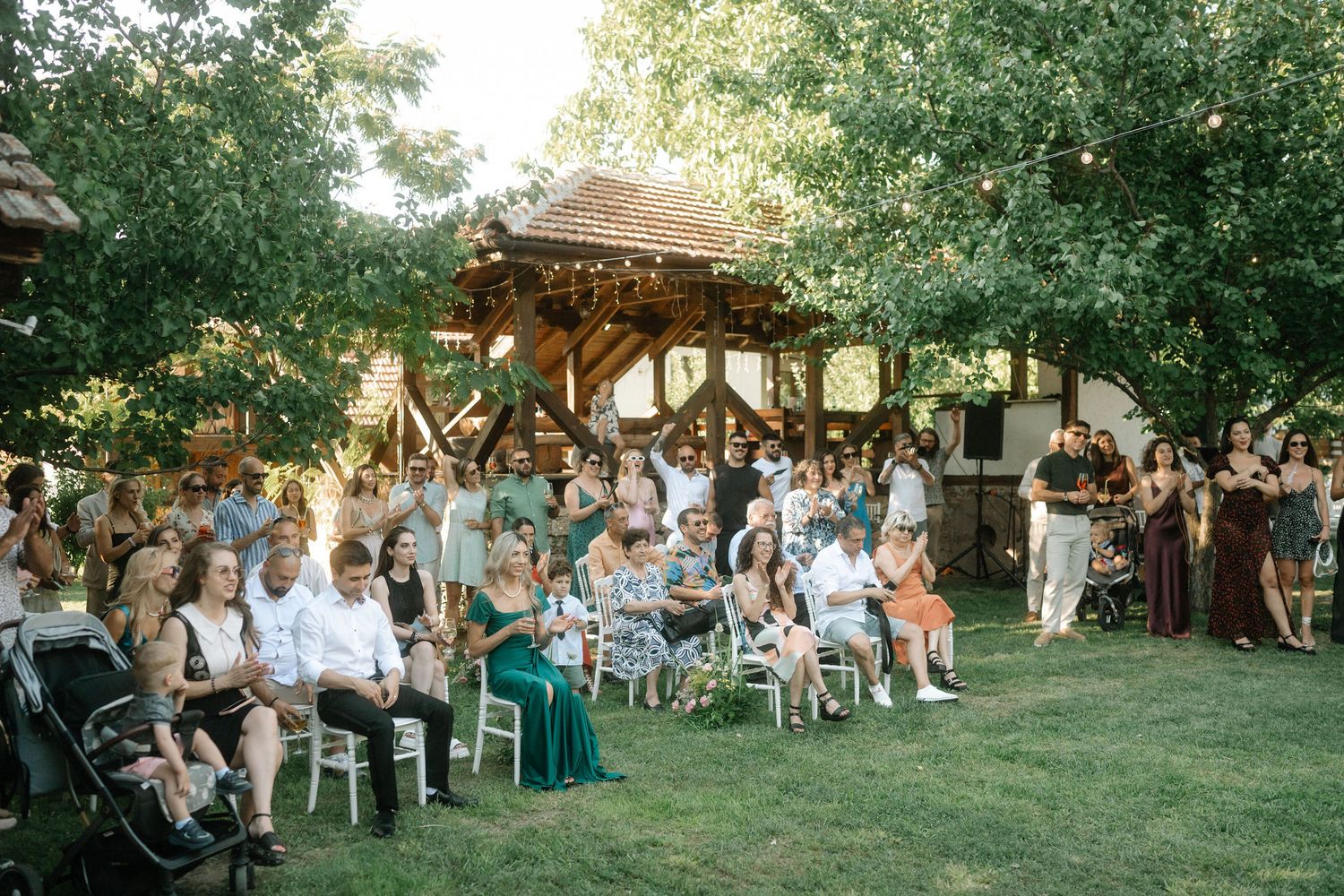 Wedding guests seated in white chairs on a lawn during an outdoor summer ceremony at dusk.