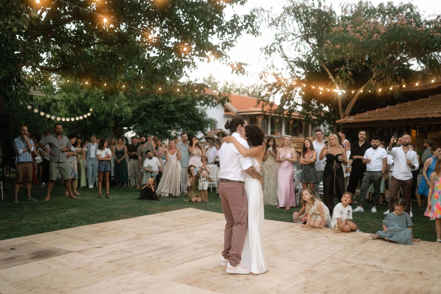 A couple shares their first dance on an outdoor dance floor under string lights while guests watch at a sunset wedding reception.