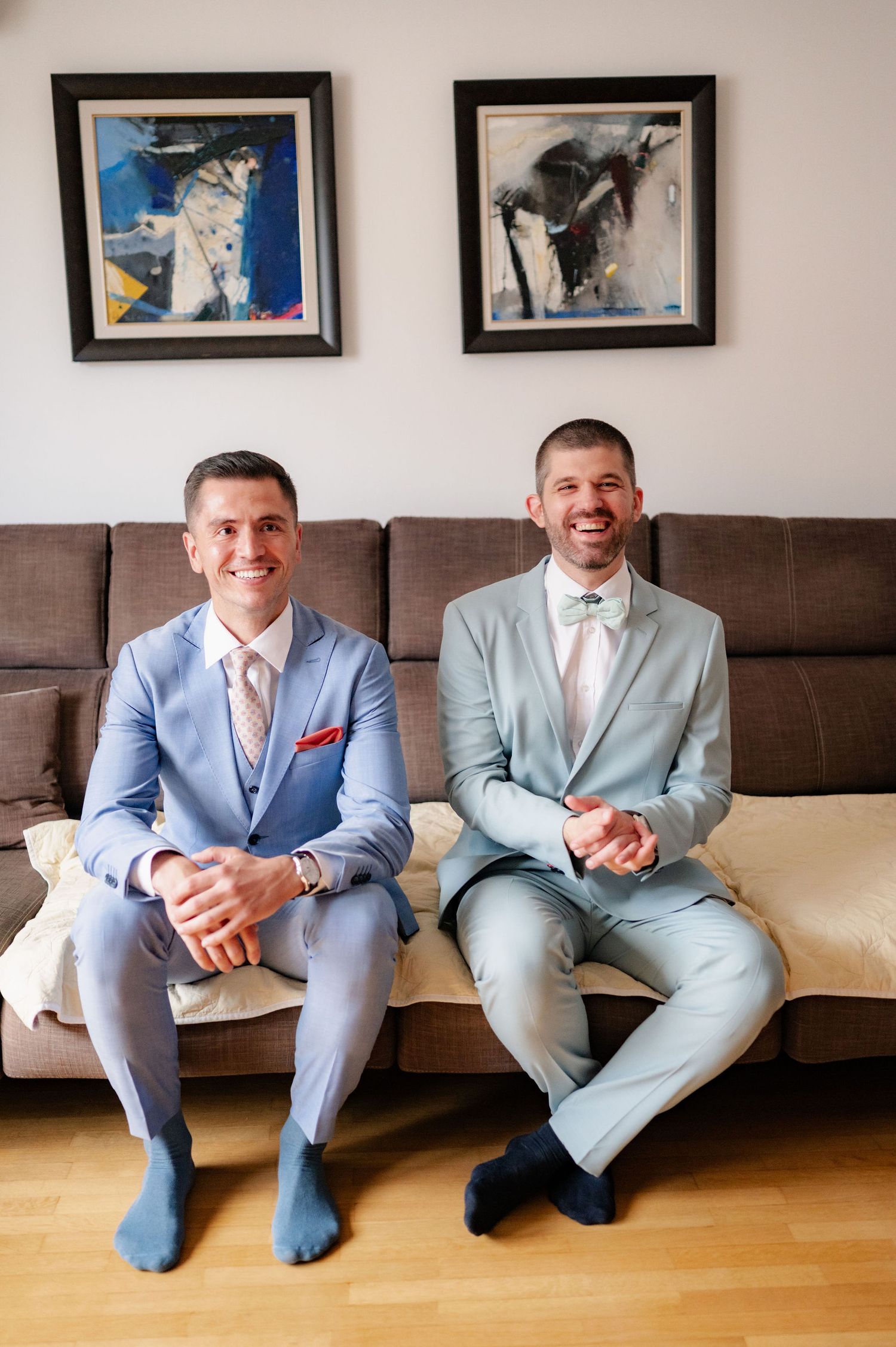 Two people in light colored suits sit together on a brown couch beneath framed abstract artwork on the wall.