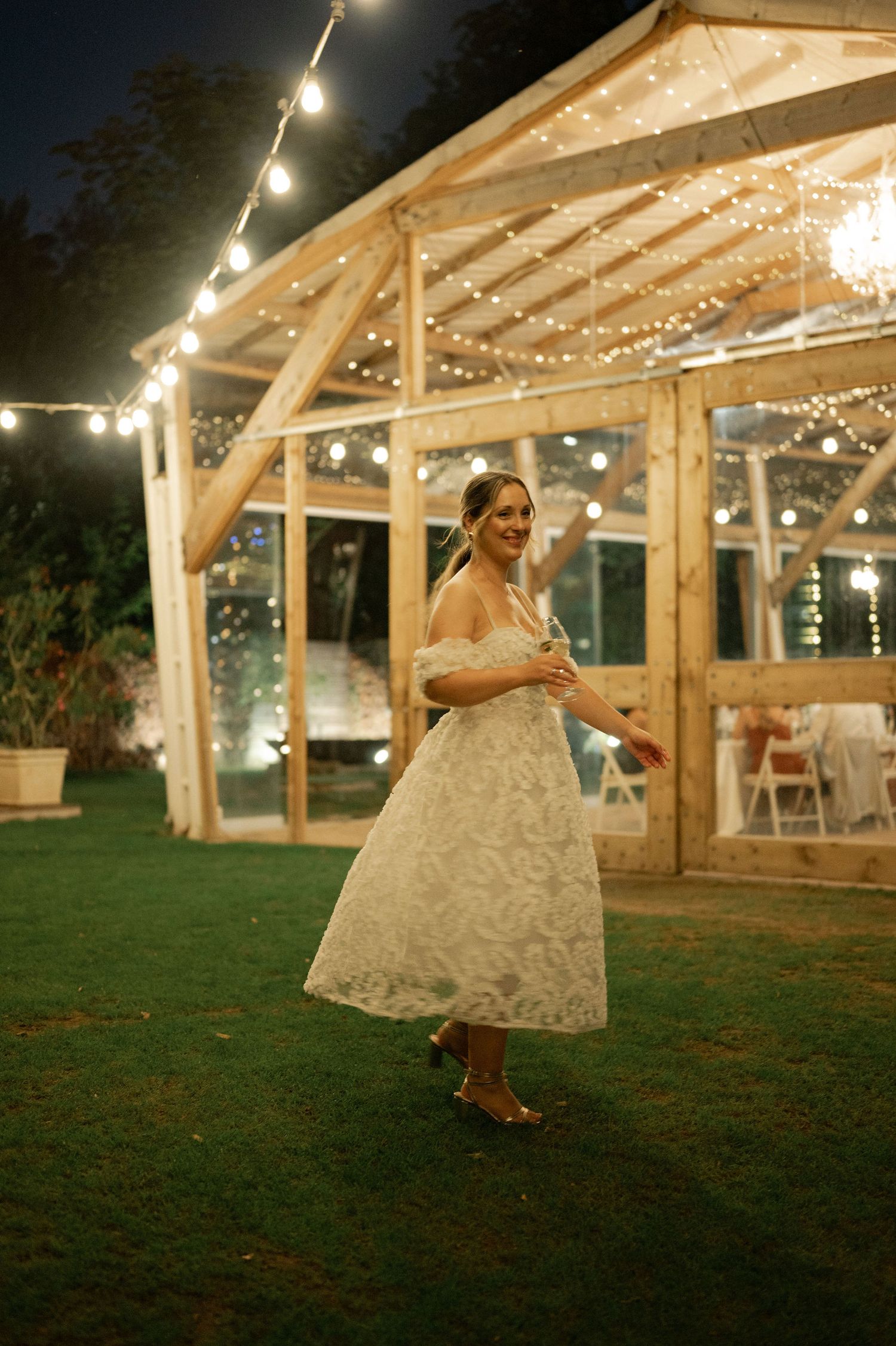 A wedding reception at dusk with string lights illuminating a rustic wooden barn structure and grass lawn.