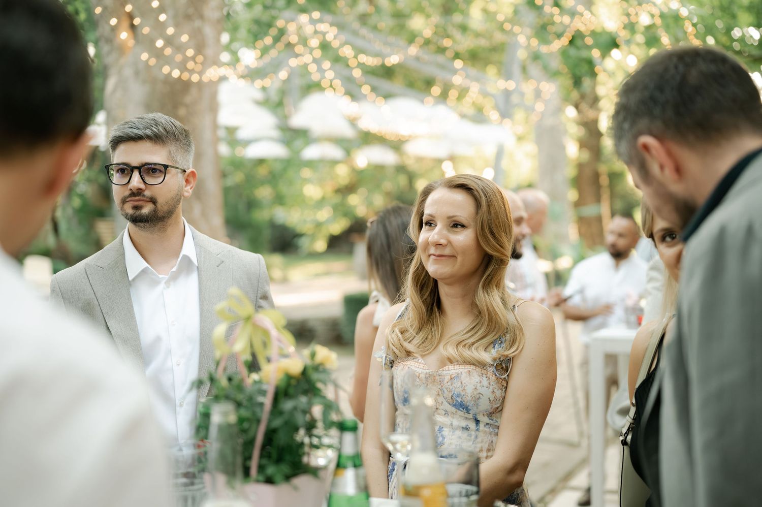 Guests mingle at an elegant outdoor garden party with twinkling lights and greenery in the background.
