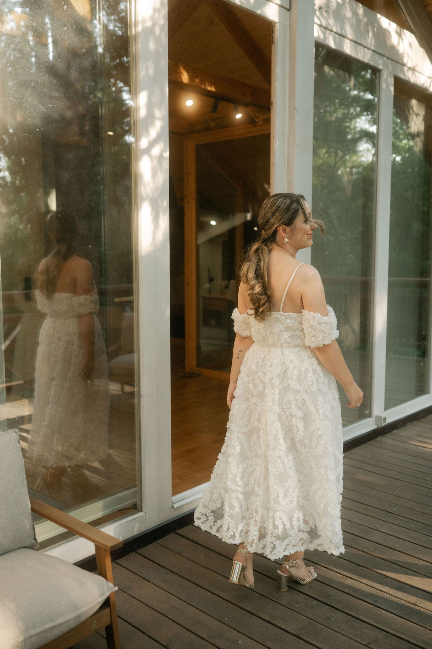 Elegant off-shoulder white lace wedding dress shown from behind on a wooden deck with glass windows reflecting natural light.