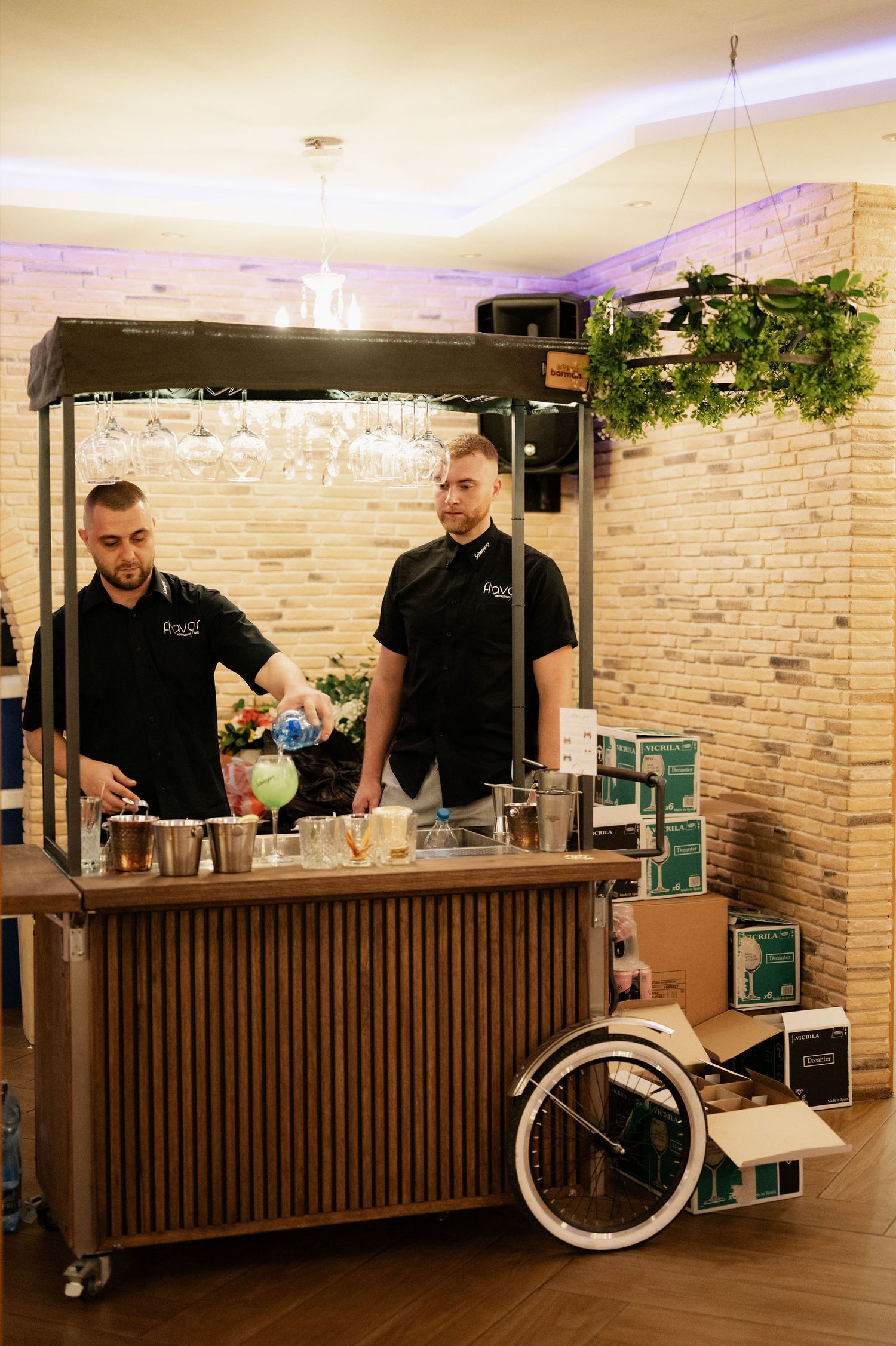 Professional bartenders prepare cocktails at a wooden slatted bar counter with decorative lighting and brick walls.