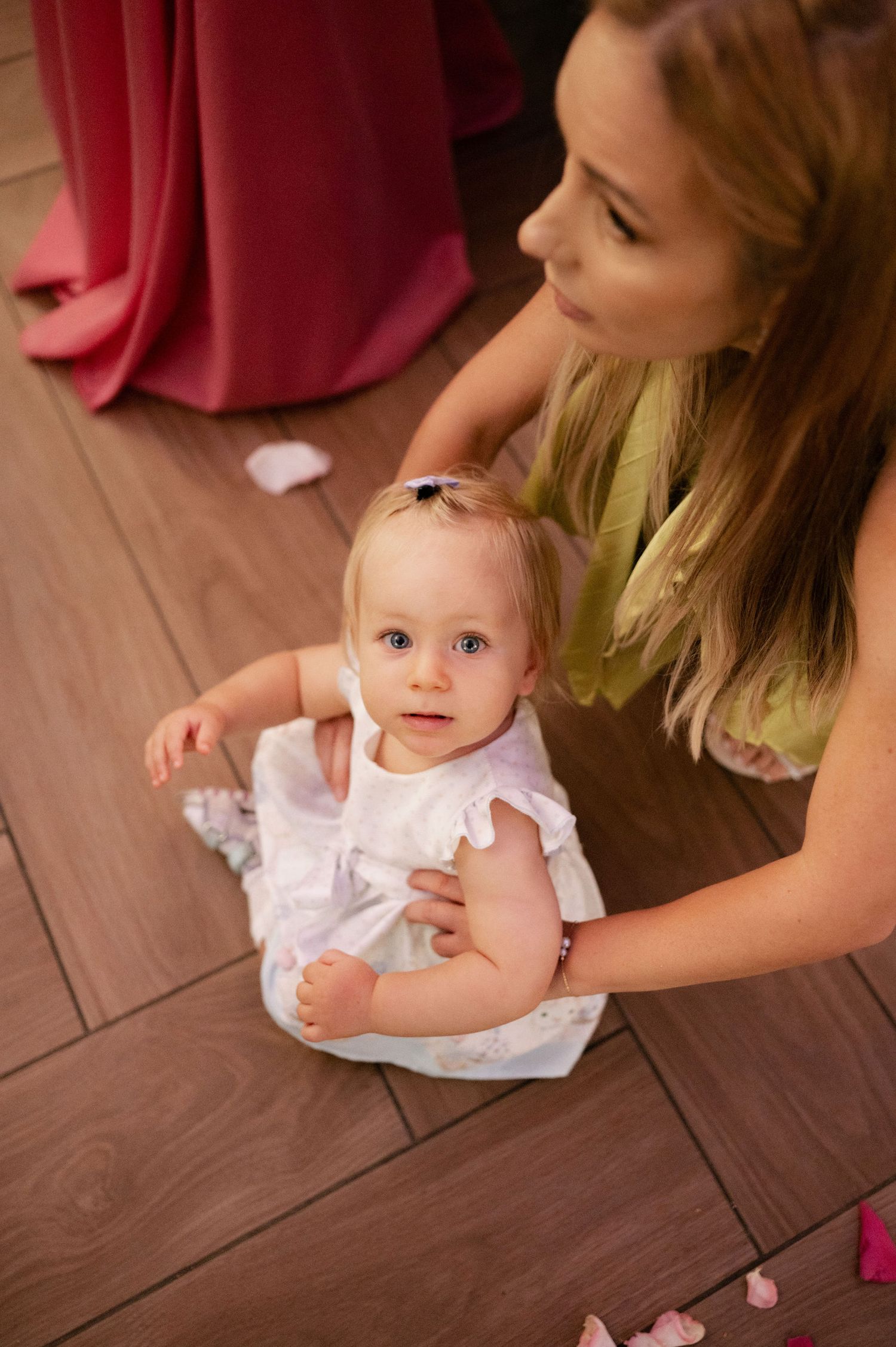 A baby in a white dress sits on dark wooden flooring while being supported by someone nearby.