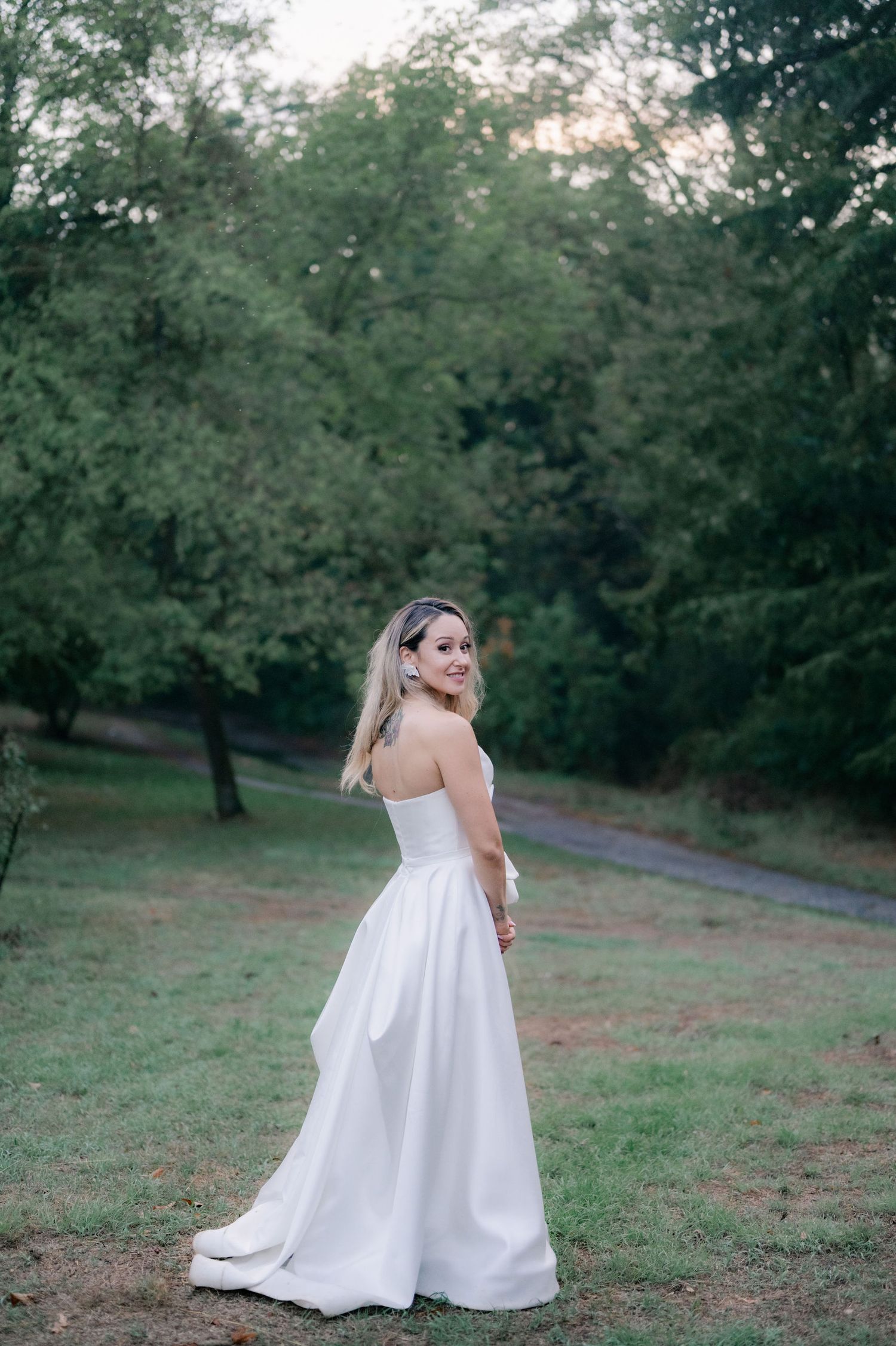 A person in a flowing white wedding dress stands gracefully in a park setting surrounded by green trees.