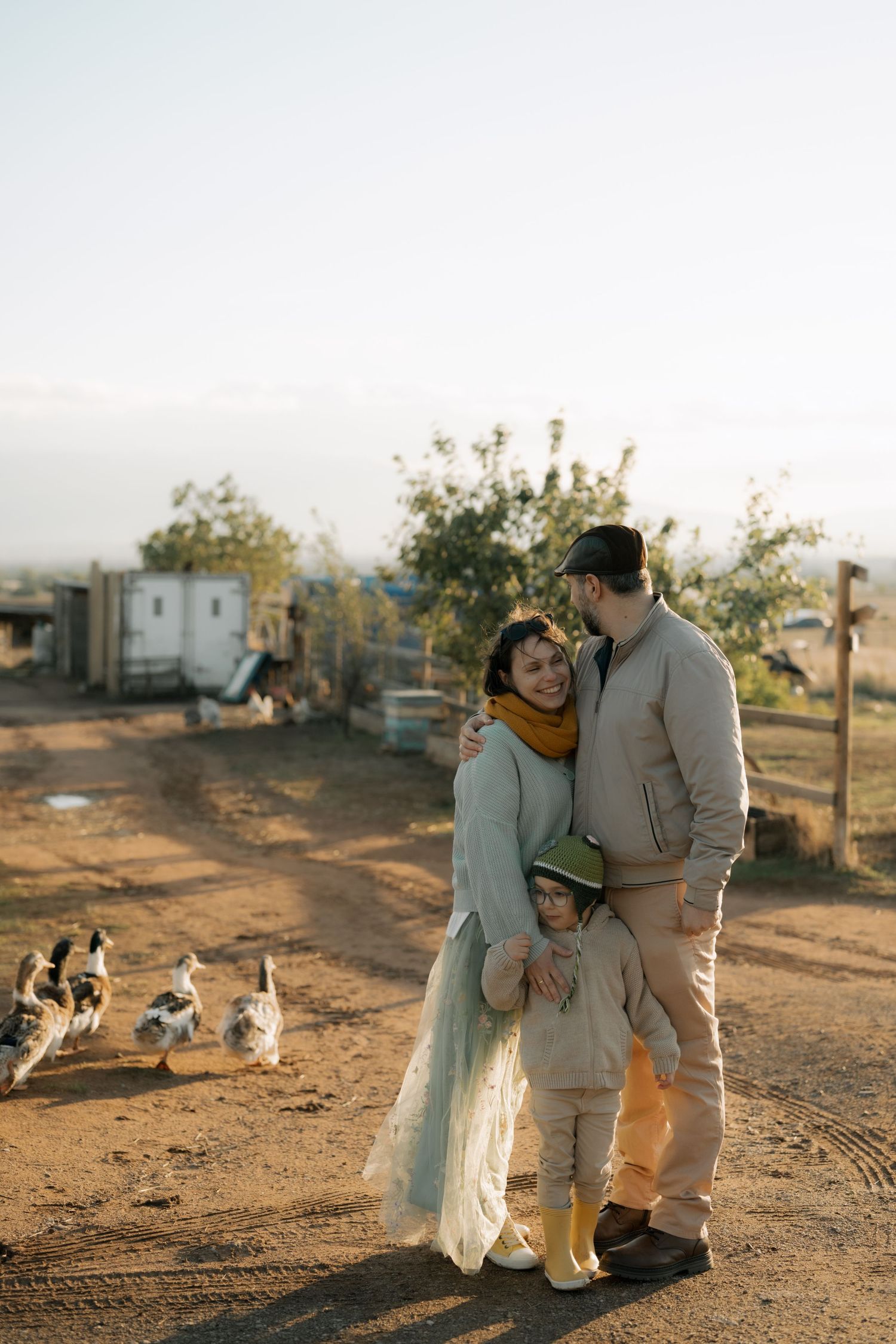 A family embraces while standing on a dirt path at sunset with chickens roaming in the background.