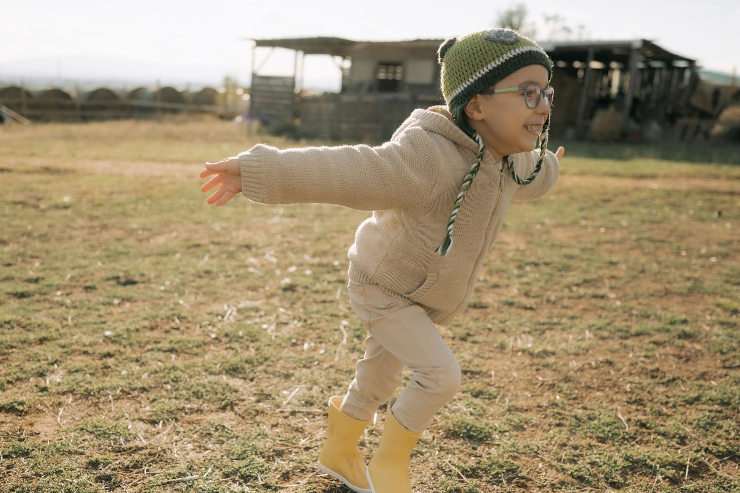 A child wearing a beige sweater and glasses plays outdoors in a field on a sunny day.