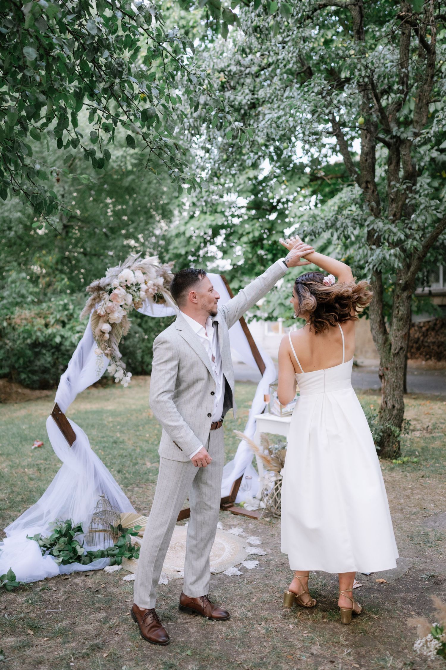 A wedding party dances and poses playfully outdoors in a wooded setting wearing white and beige attire.