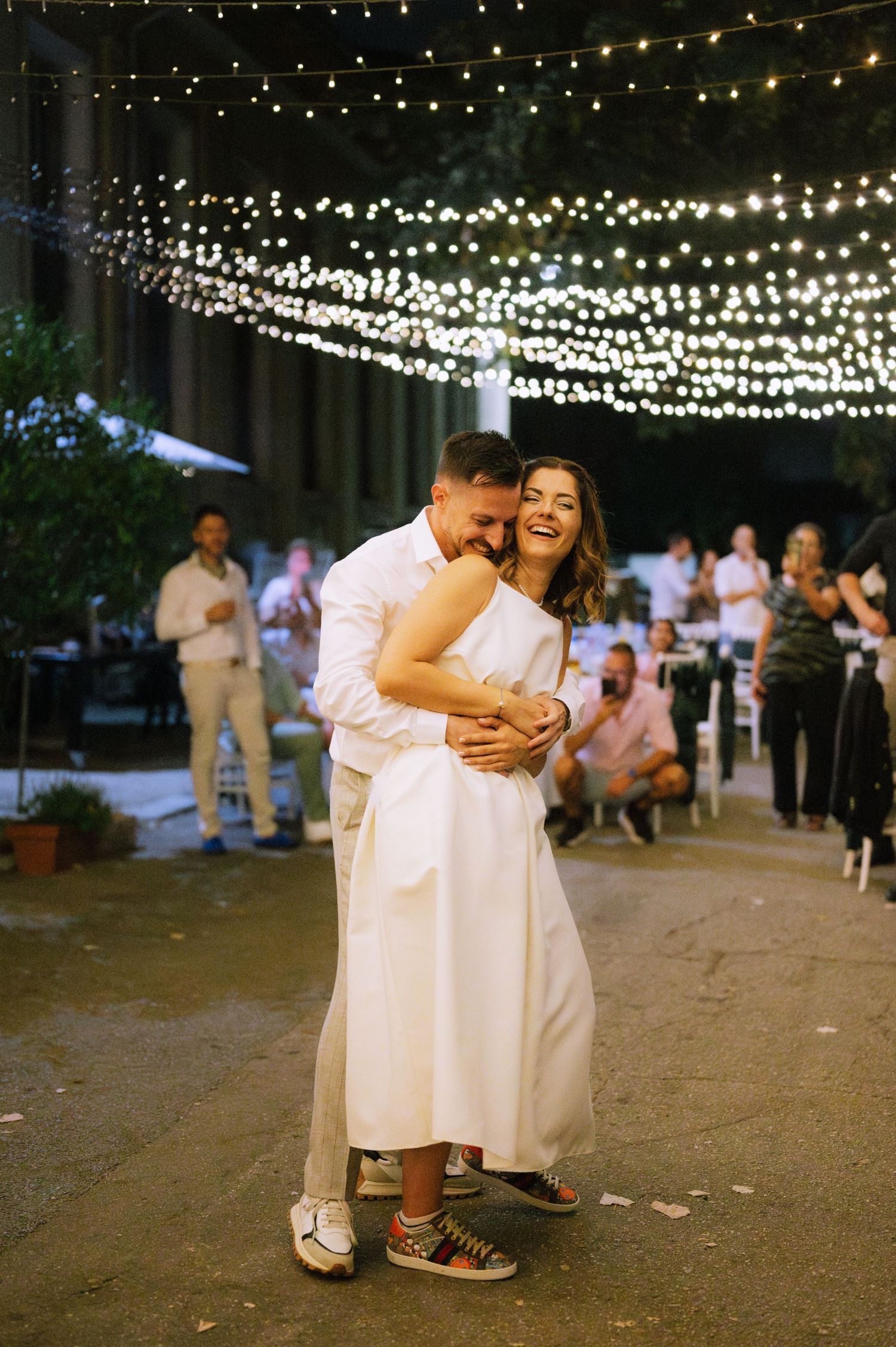 String lights illuminate an outdoor dance floor where people in white outfits dance together at a festive celebration.